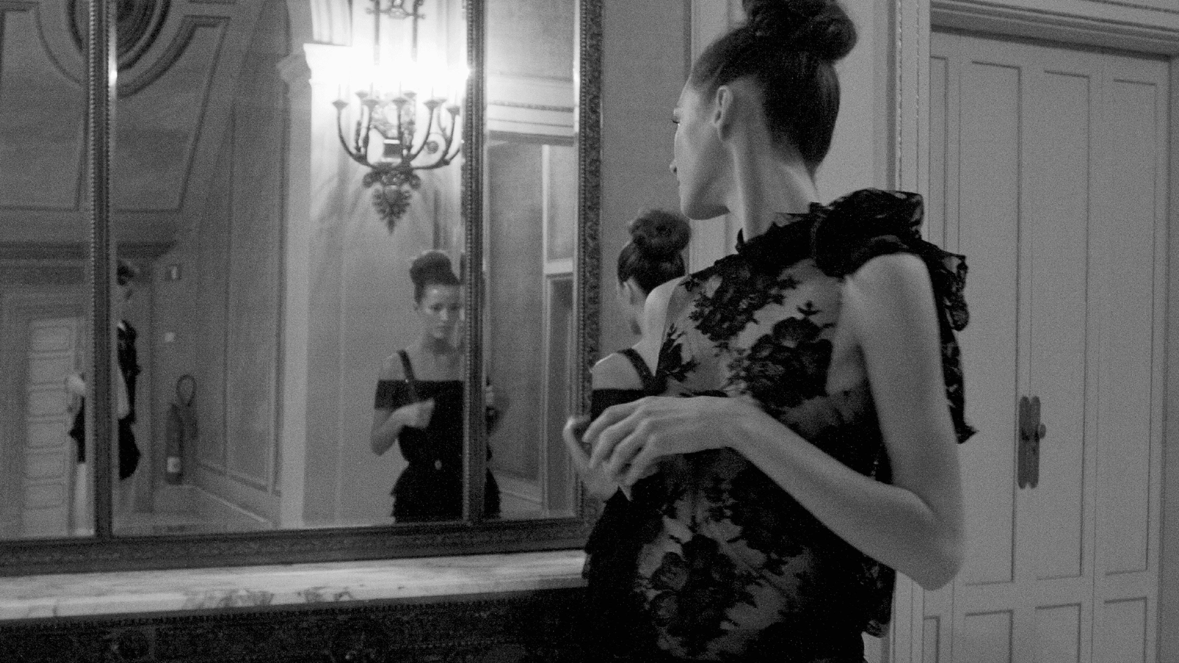 A woman in a sophisticated black evening gown admires her reflection in a grand mirror, capturing a moment of classic elegance.