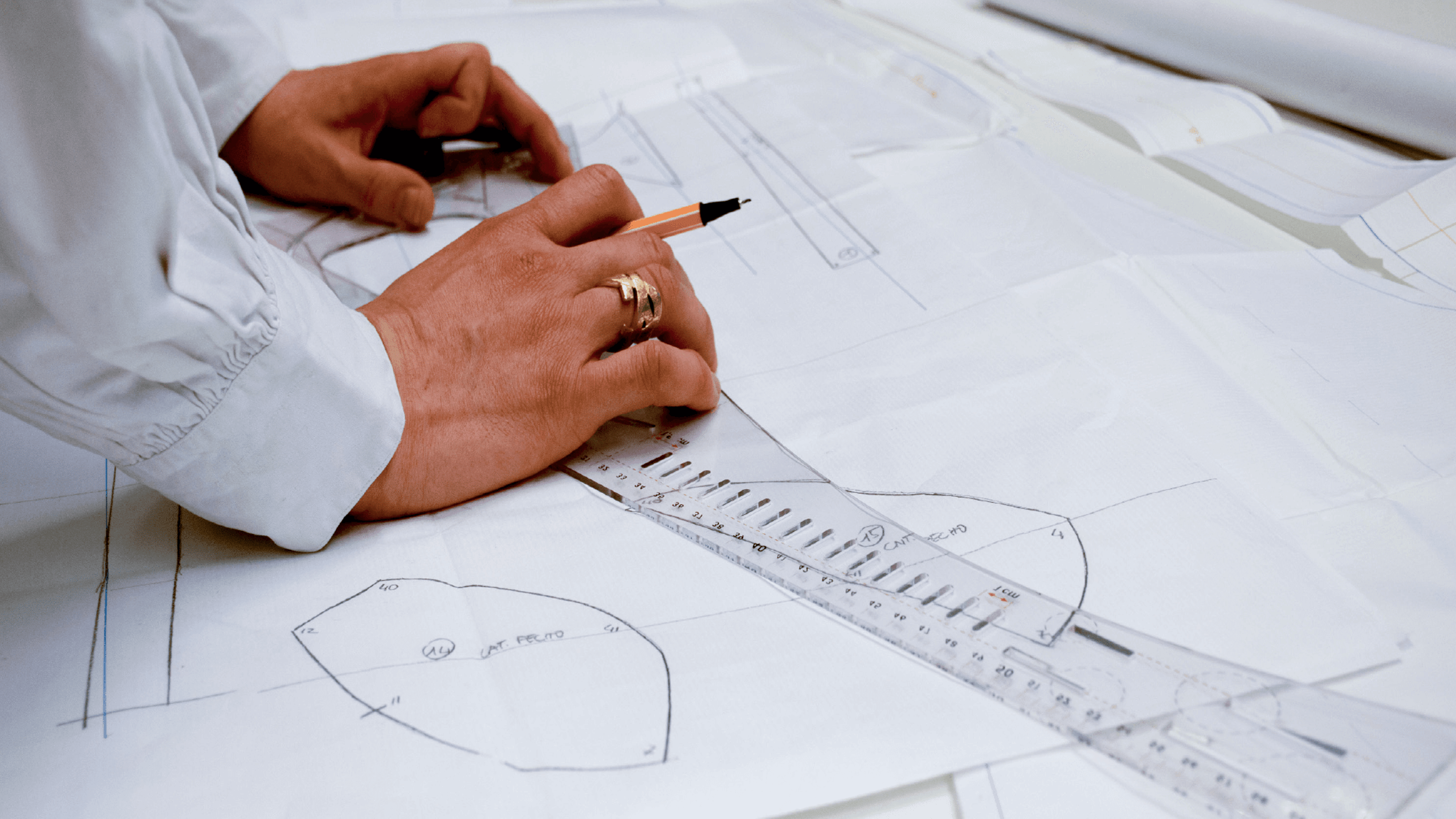 Primer plano de las manos de un diseñador utilizando una regla y un lápiz para diseñar un patrón de ropa, destacando la meticulosa planificación de la prenda.

