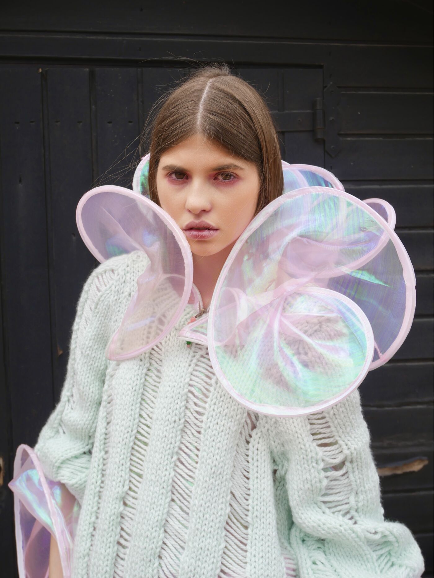 A young woman is featured wearing a light-colored knitted sweater adorned with a voluminous, iridescent collar. The background is dark.