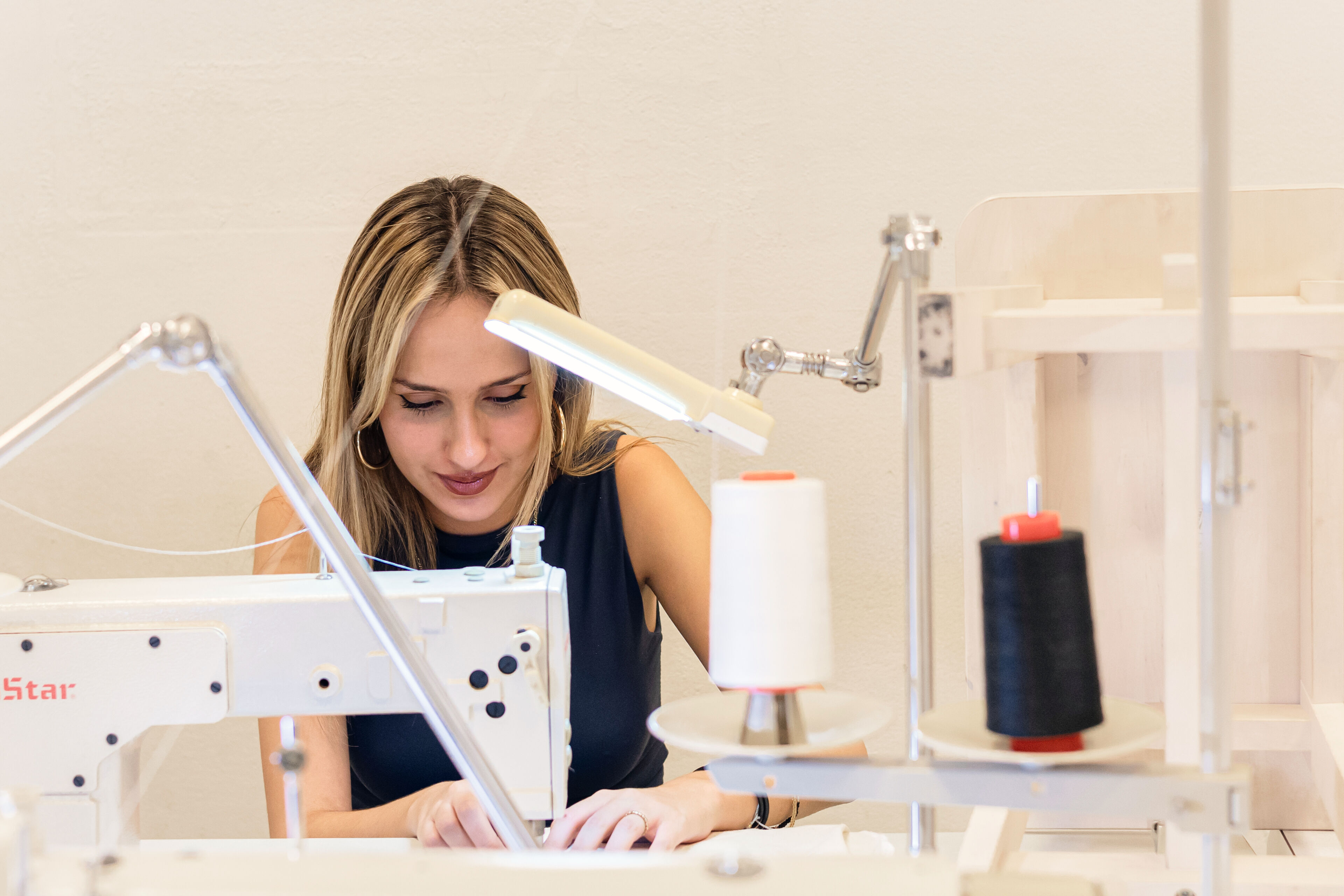 Una dona utilitzant atentament una màquina de cosir professional, amb carretells de fil i una llum de tasca a l'espai de treball.

