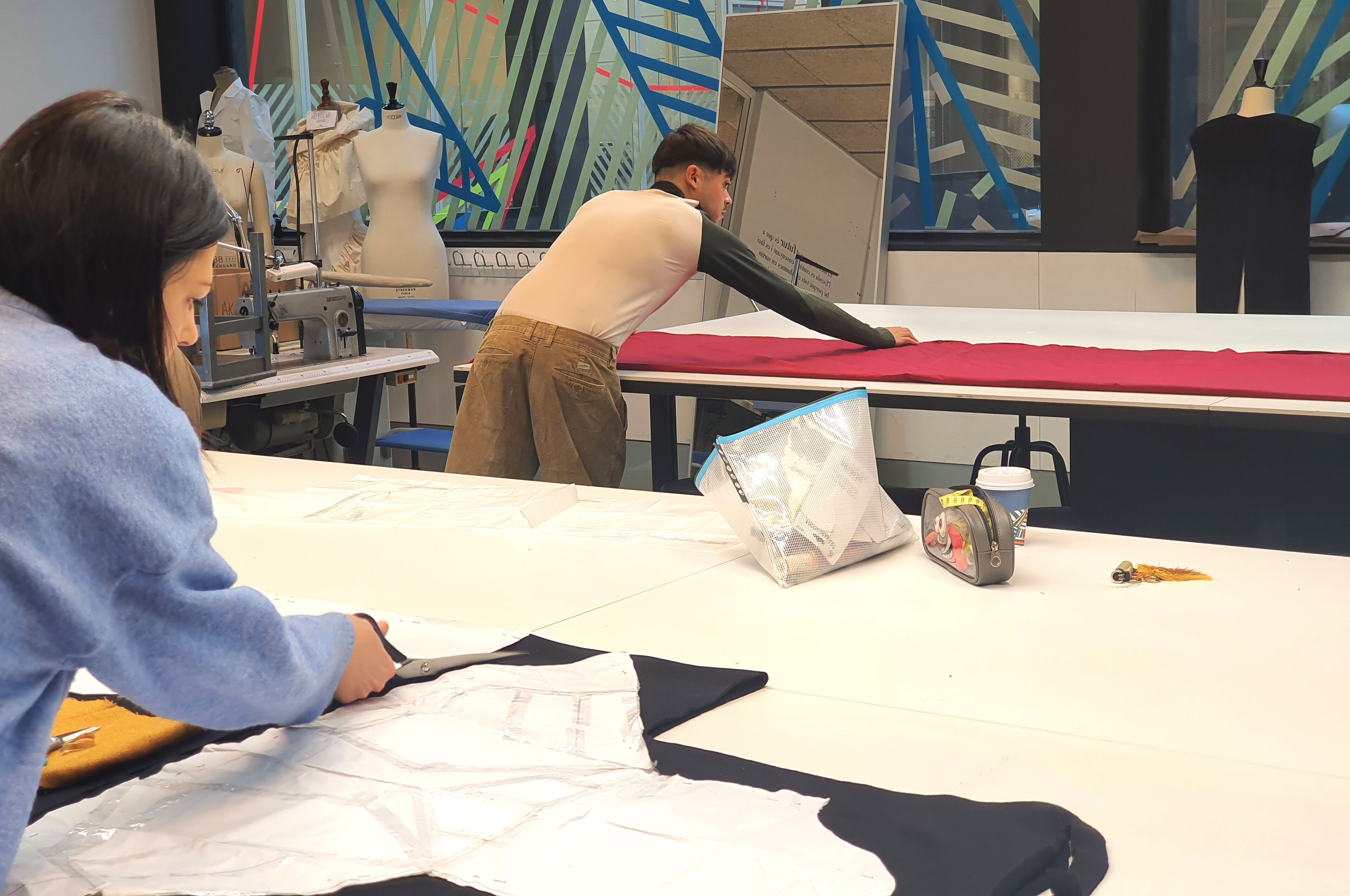 Estudiantes de diseño de moda están ocupados cortando y preparando tela en grandes mesas en un estudio de diseño iluminado.

