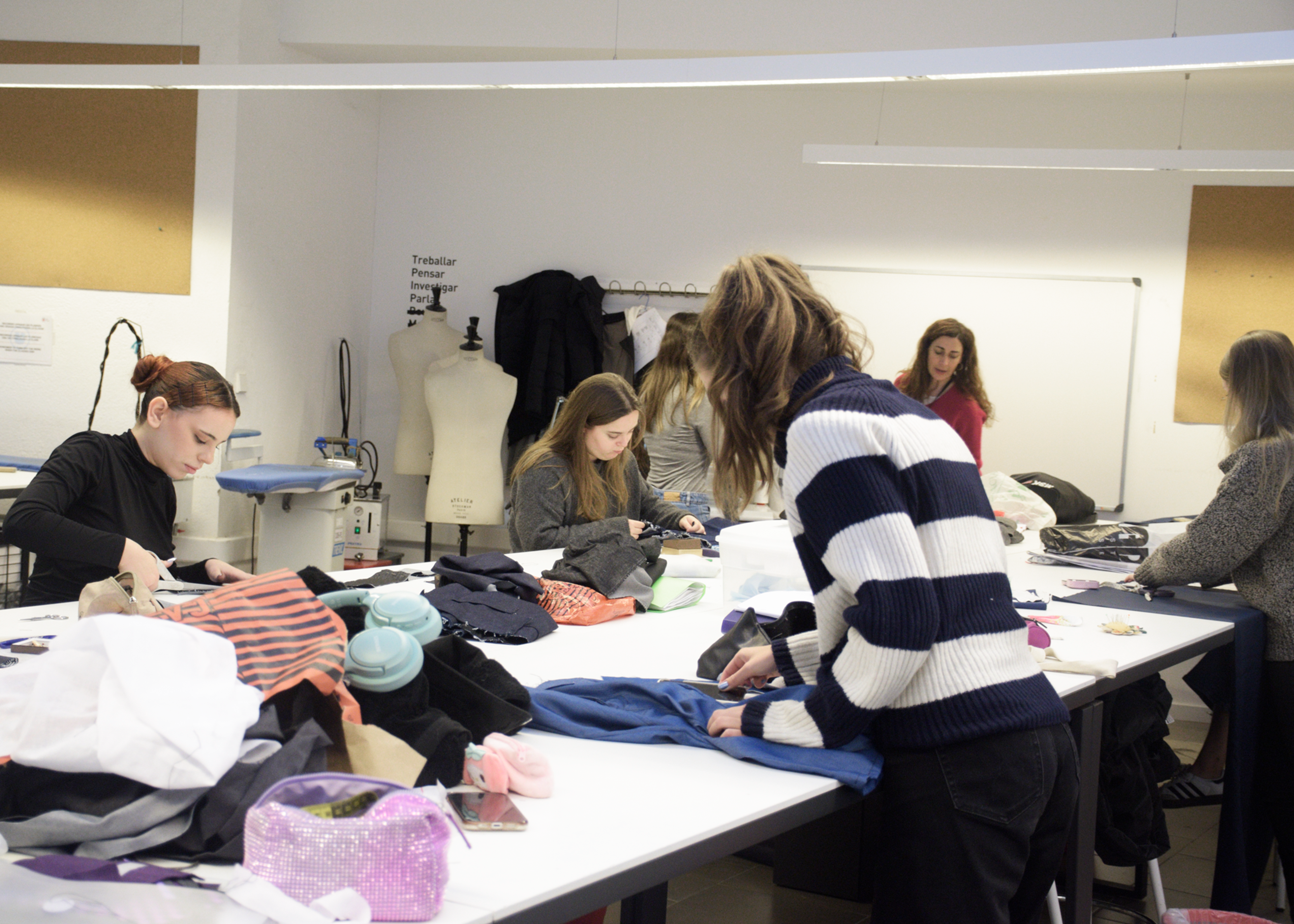 Estudiantes de moda trabajan diligentemente en varias prendas en una mesa de estudio concurrida, rodeados de tela y herramientas de diseño.

