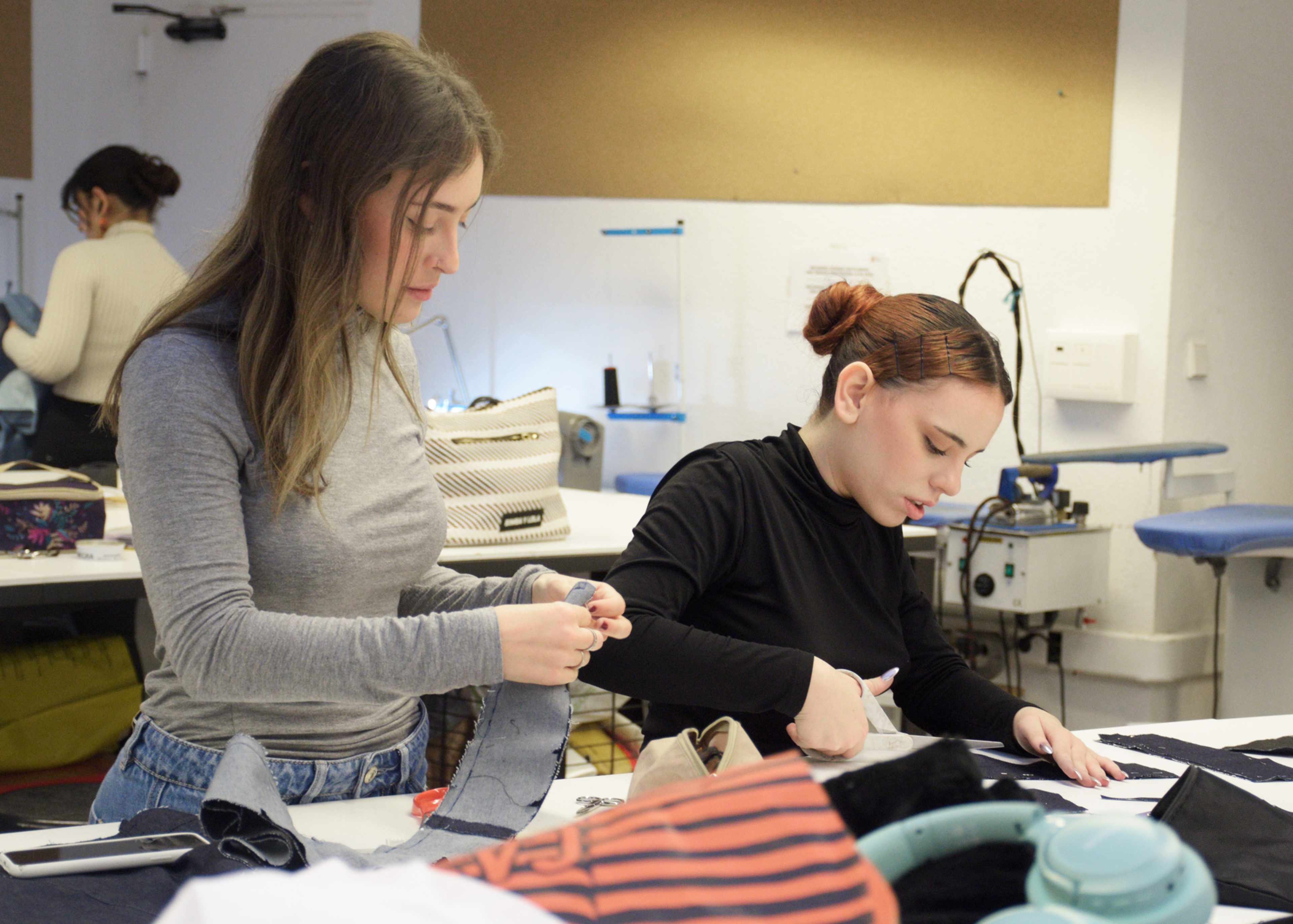 Estudiantes de moda concentrados cosiendo a mano y ensamblando piezas en un taller, rodeados de tela y equipo de costura.

