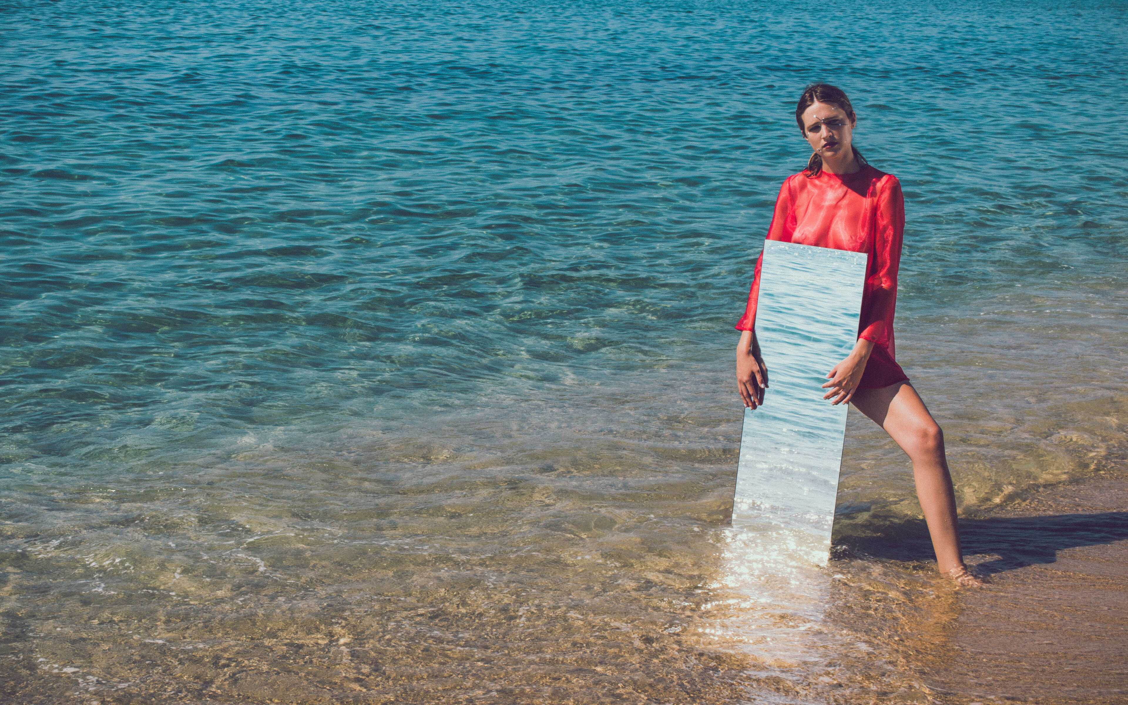 Una dona amb blusa vermella està al mar, aguantant un mirall que reflecteix l'aigua, creant una intersecció surrealista entre imatge i realitat.