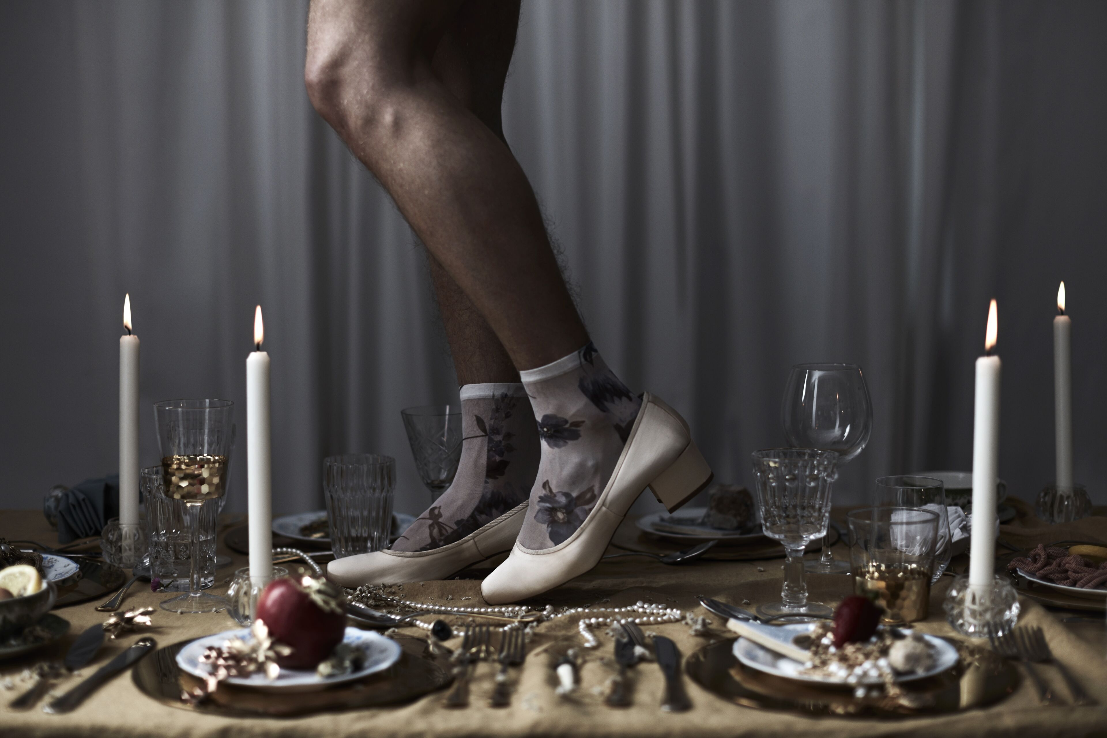 Piernas de una persona con calcetines florales y tacones sobre una mesa de cena con platos y velas, estableciendo un tono caprichoso.