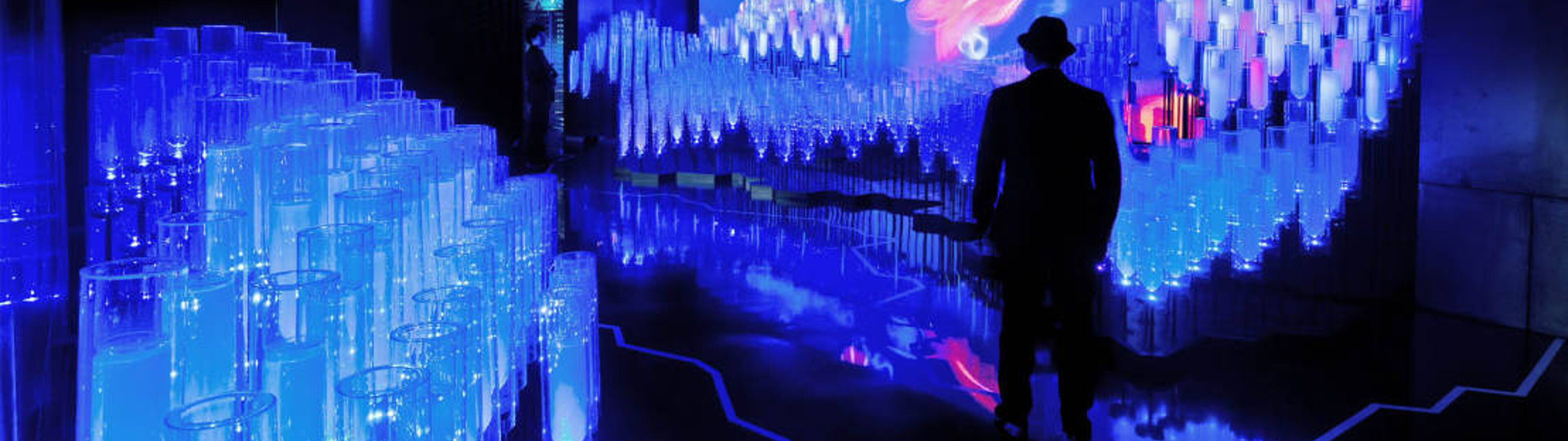 Persona parada en medio de una llamativa instalación de arte luminiscente azul con patrones de luz dinámicos proyectados en la pared.