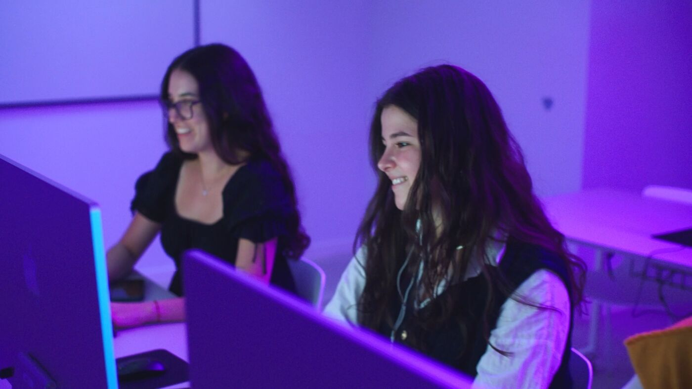 Two young women work on computers in a classroom with vibrant blue-purple lighting. They are focused and smiling, reflecting enthusiasm and engagement in a creative digital environment.