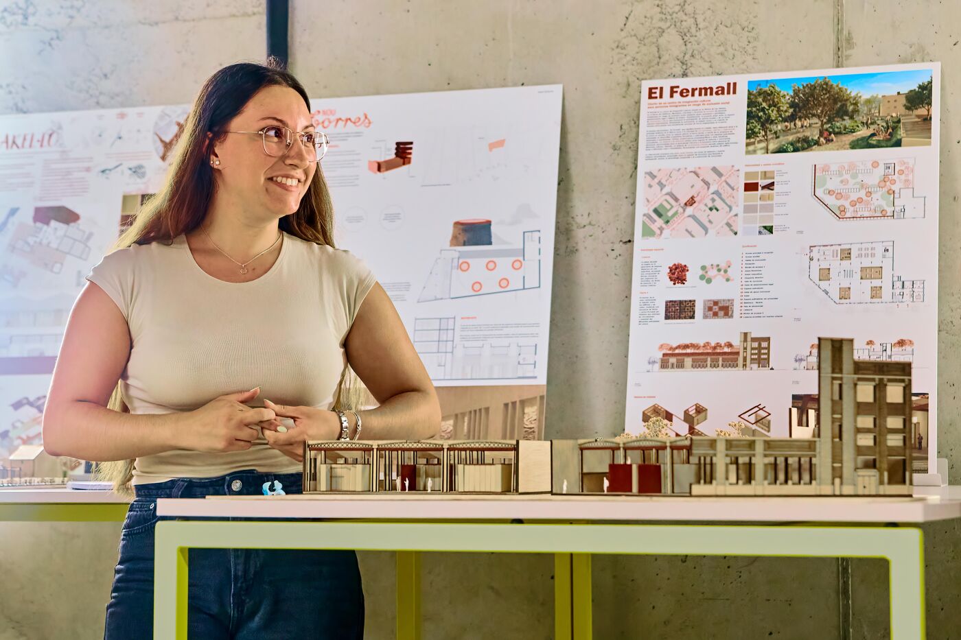 A woman stands in front of architectural models and presentation boards, with one board featuring the text "El Fermall". She appears to be presenting her work.