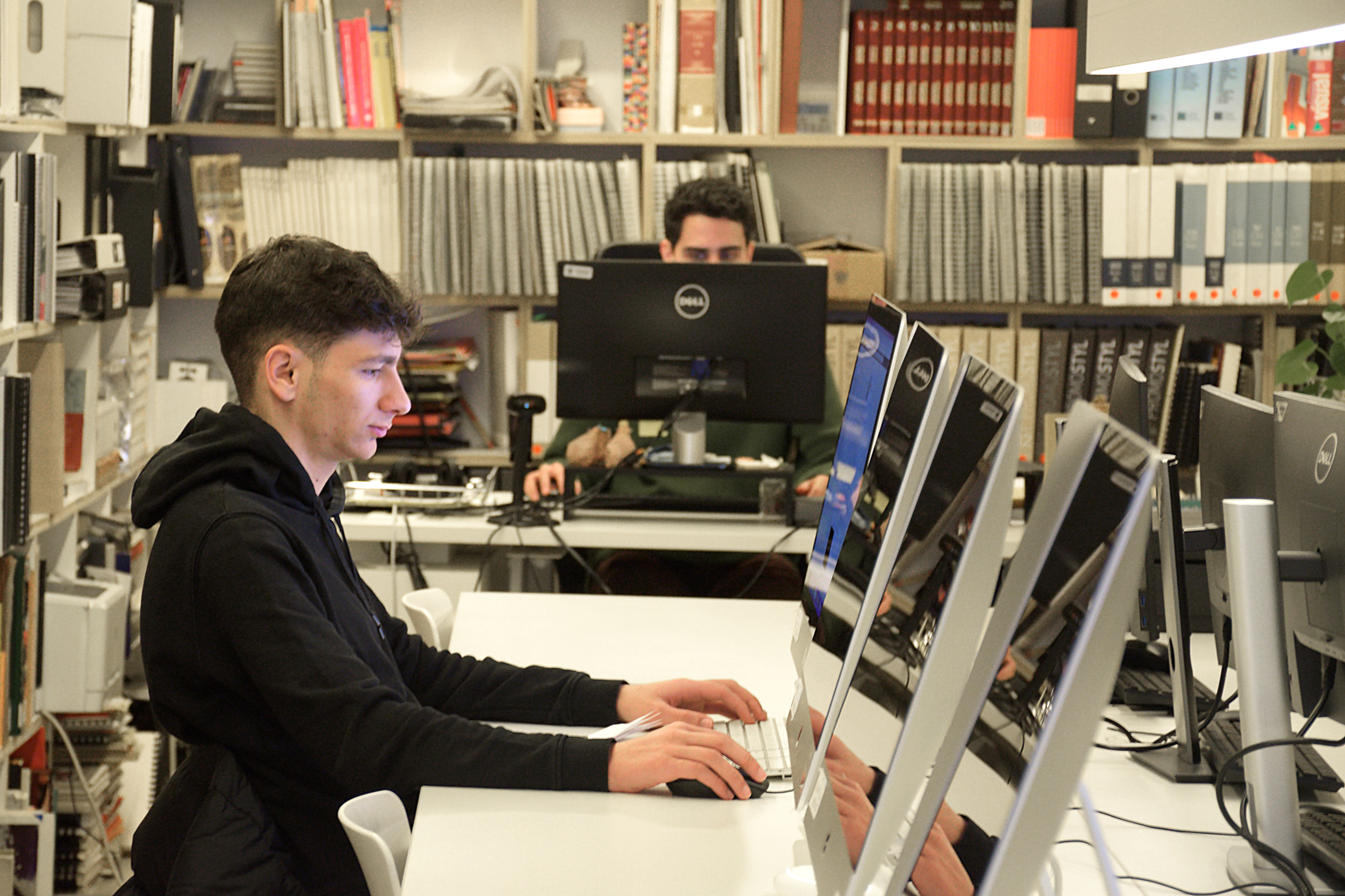 A focused individual typing on a desktop computer in a modern library with books and another person in the background.