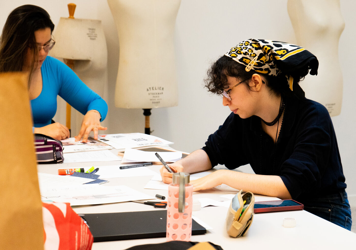 The image shows a group of people sitting around a table, working on various tasks. There are multiple cups, a bottle, and a handbag on the table, suggesting that the individuals are engaged in a casual or informal setting. The presence of a handbag indicates that the people might be in a public space or a cafe, where they can comfortably work on their projects. The group appears to be focused on their tasks, which could include drawing, writing, or discussing ideas.