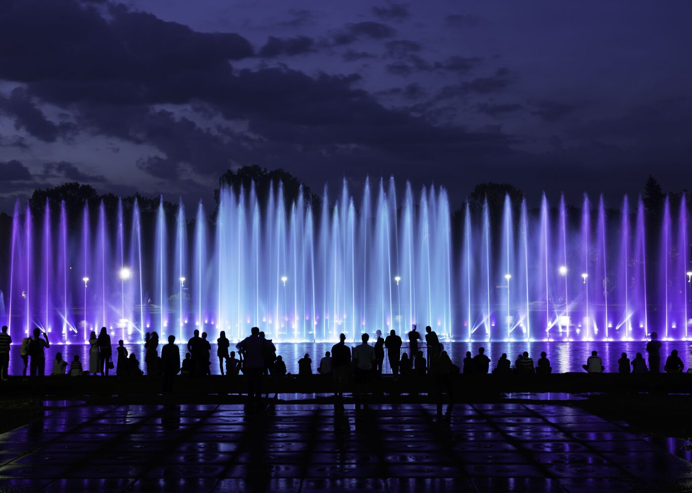 Spectators gather to watch a vibrant fountain display with beams of blue and purple light piercing through the mist against a dusky sky.