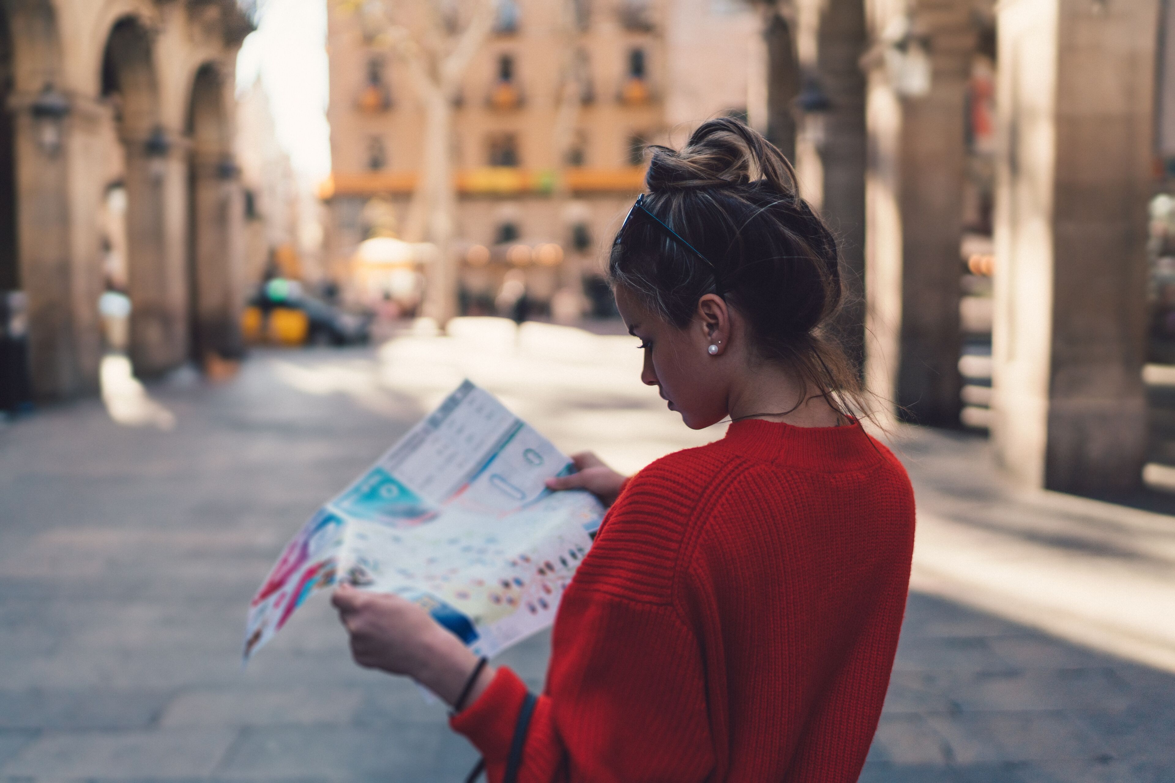 Une jeune femme en pull rouge vif étudie attentivement une carte, debout dans une rue pavée avec une architecture historique floue en arrière-plan.