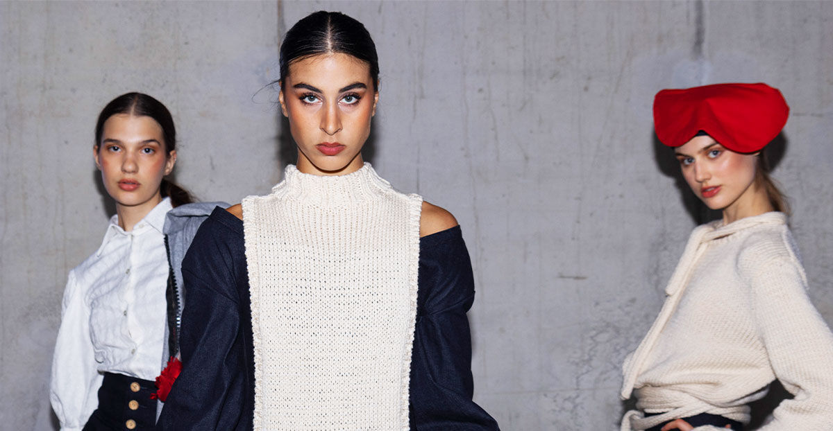 The image shows three models posing in front of a gray wall. The model in the center wears a white knit top with a navy jacket, while the other models wear white tops. One model sports a red headpiece.