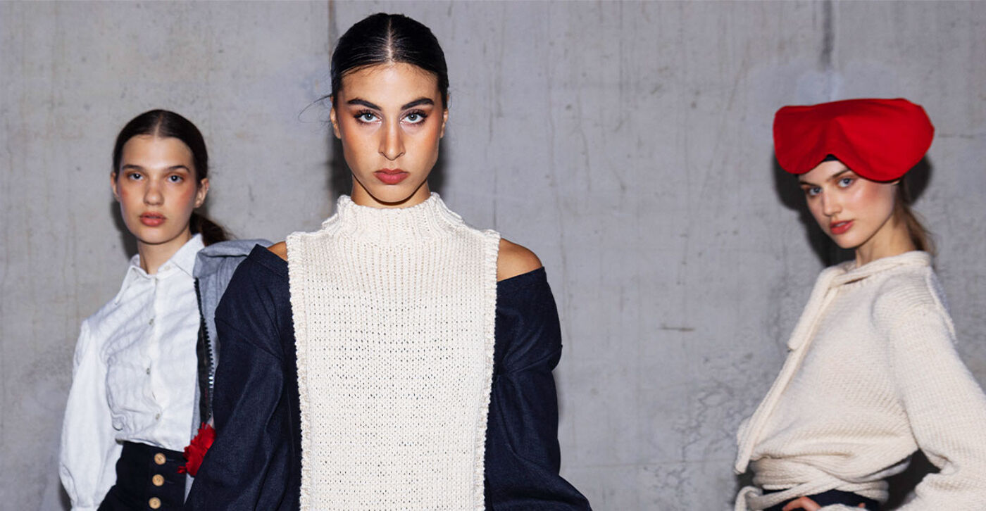 The image shows three models posing in front of a gray wall. The model in the center wears a white knit top with a navy jacket, while the other models wear white tops. One model sports a red headpiece.