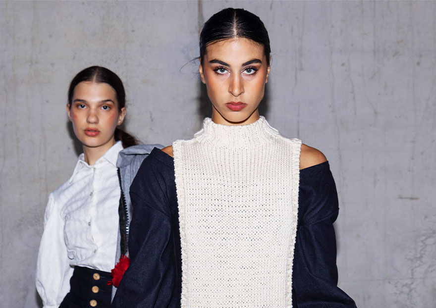 The image shows three models posing in front of a gray wall. The model in the center wears a white knit top with a navy jacket, while the other models wear white tops. One model sports a red headpiece.