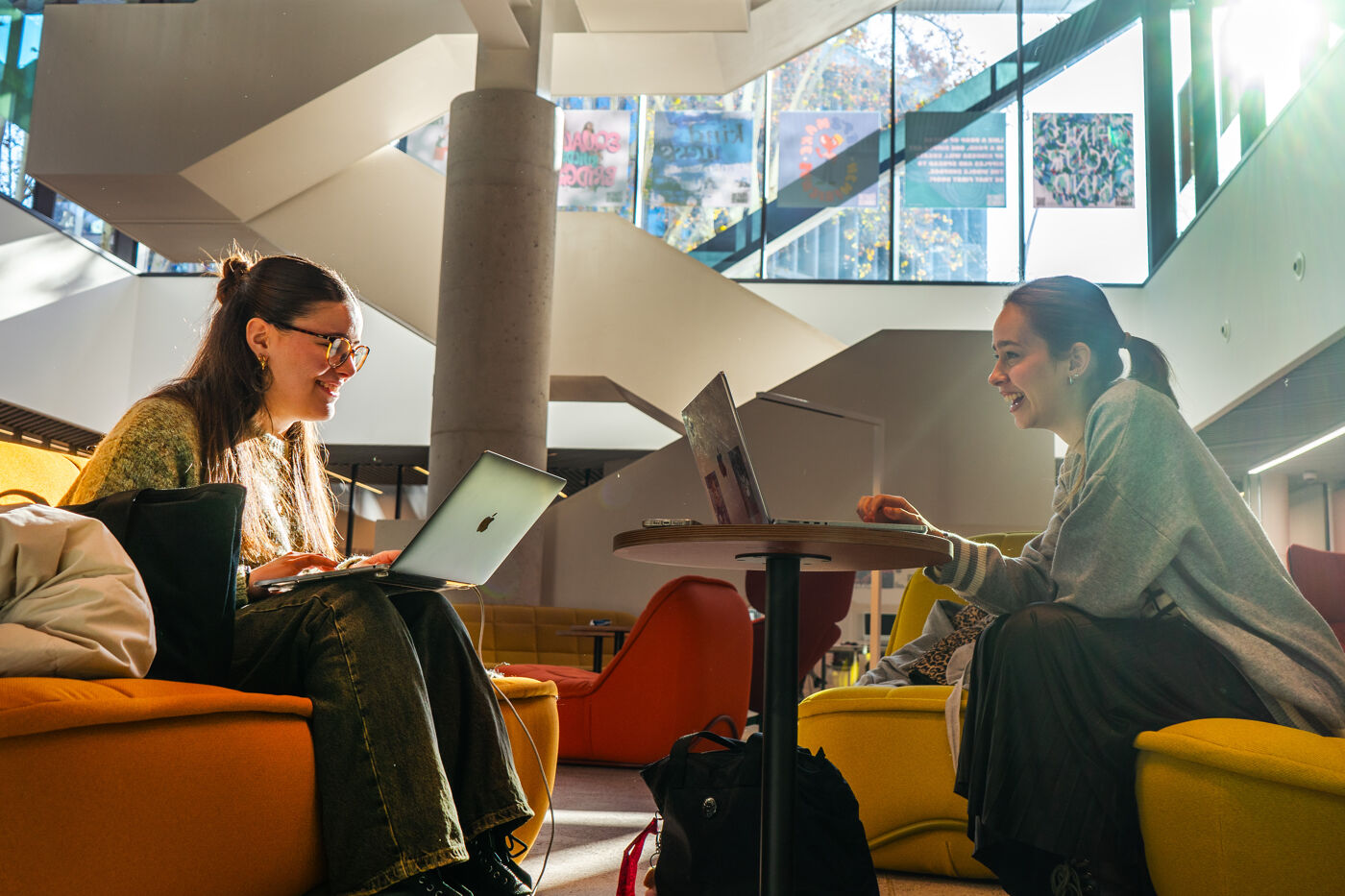 Two young women sit on colorful armchairs in a modern building's bright interior, each working on their own laptop.