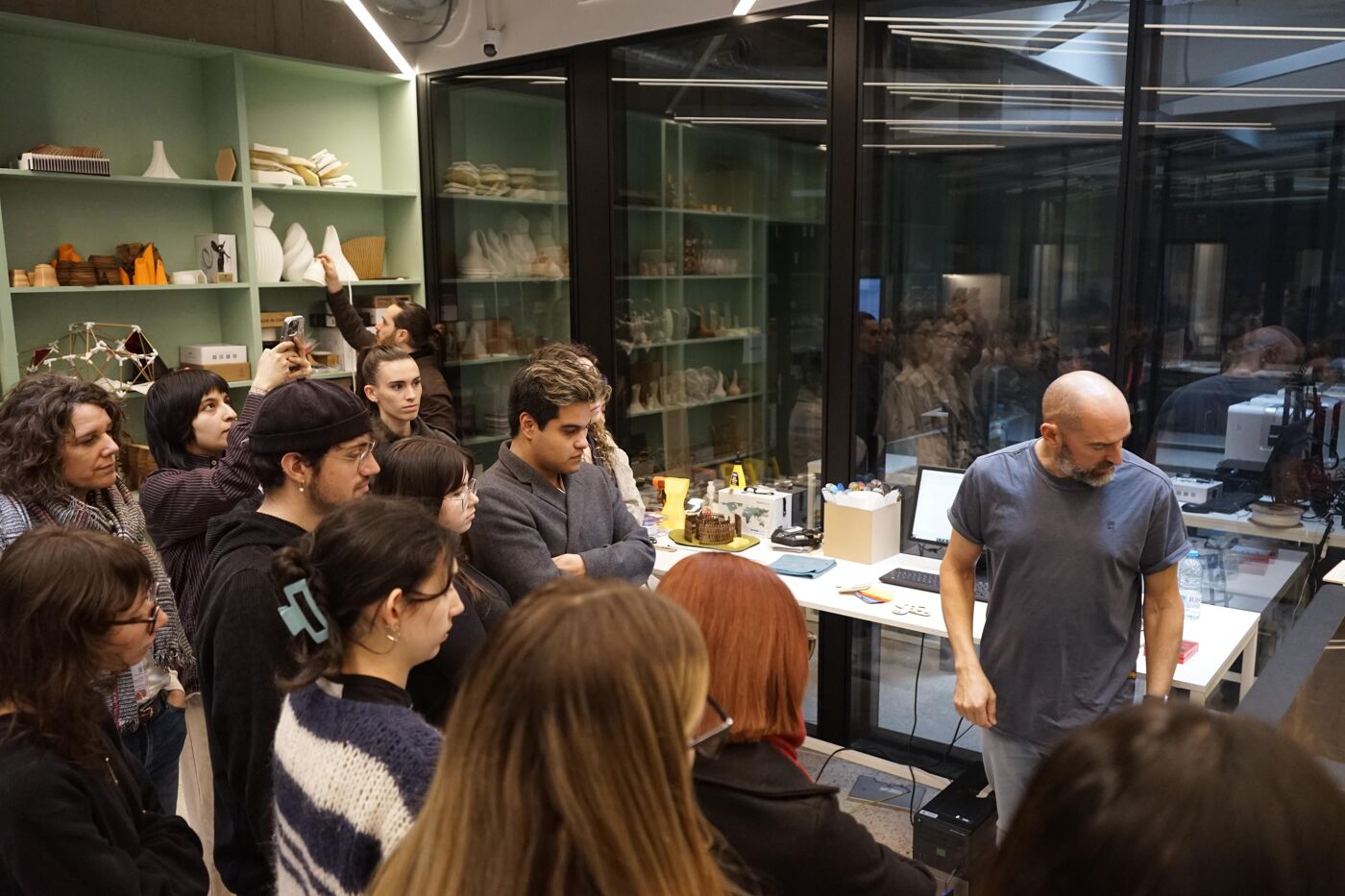 A diverse group of people, many young adults, are gathered in a brightly lit room with shelves filled with objects, attentively listening to a man in a gray t-shirt who is speaking.