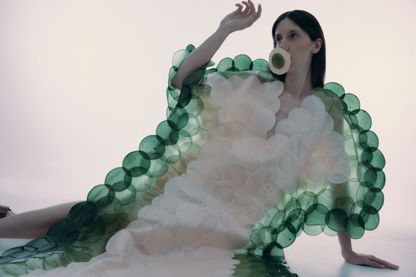 A woman in a white and green circular dress plays with a bubble gum bubble. The dress has a scale-like design, reminiscent of a sea creature.