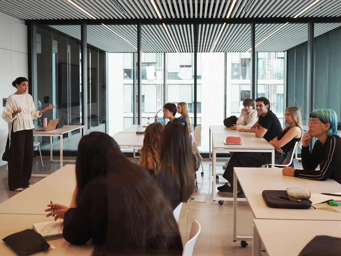 A woman is lecturing a group of students in a modern classroom with large windows overlooking an office building.