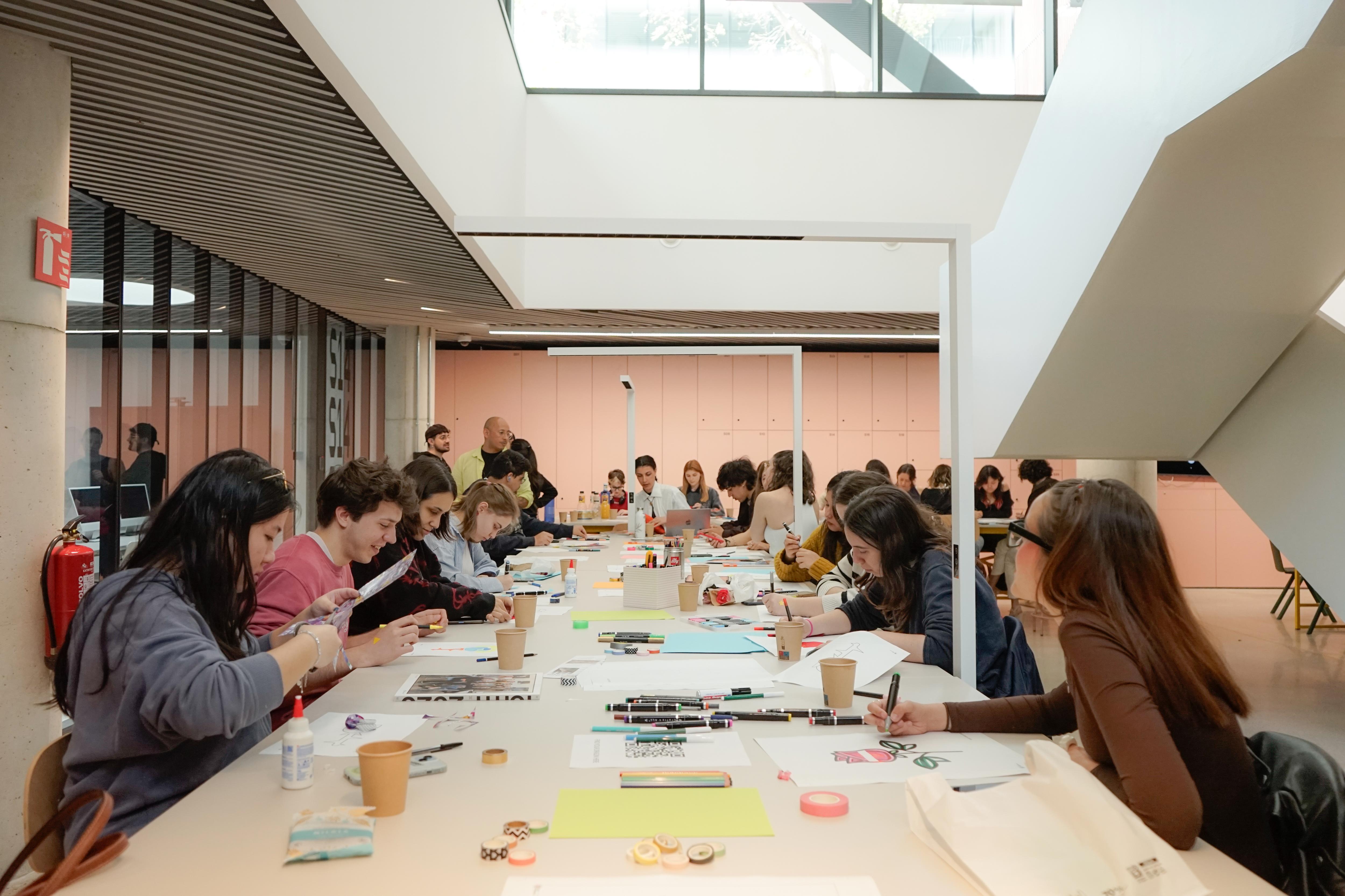 A large group of people sit at long tables in a bright, modern building, working on various projects.