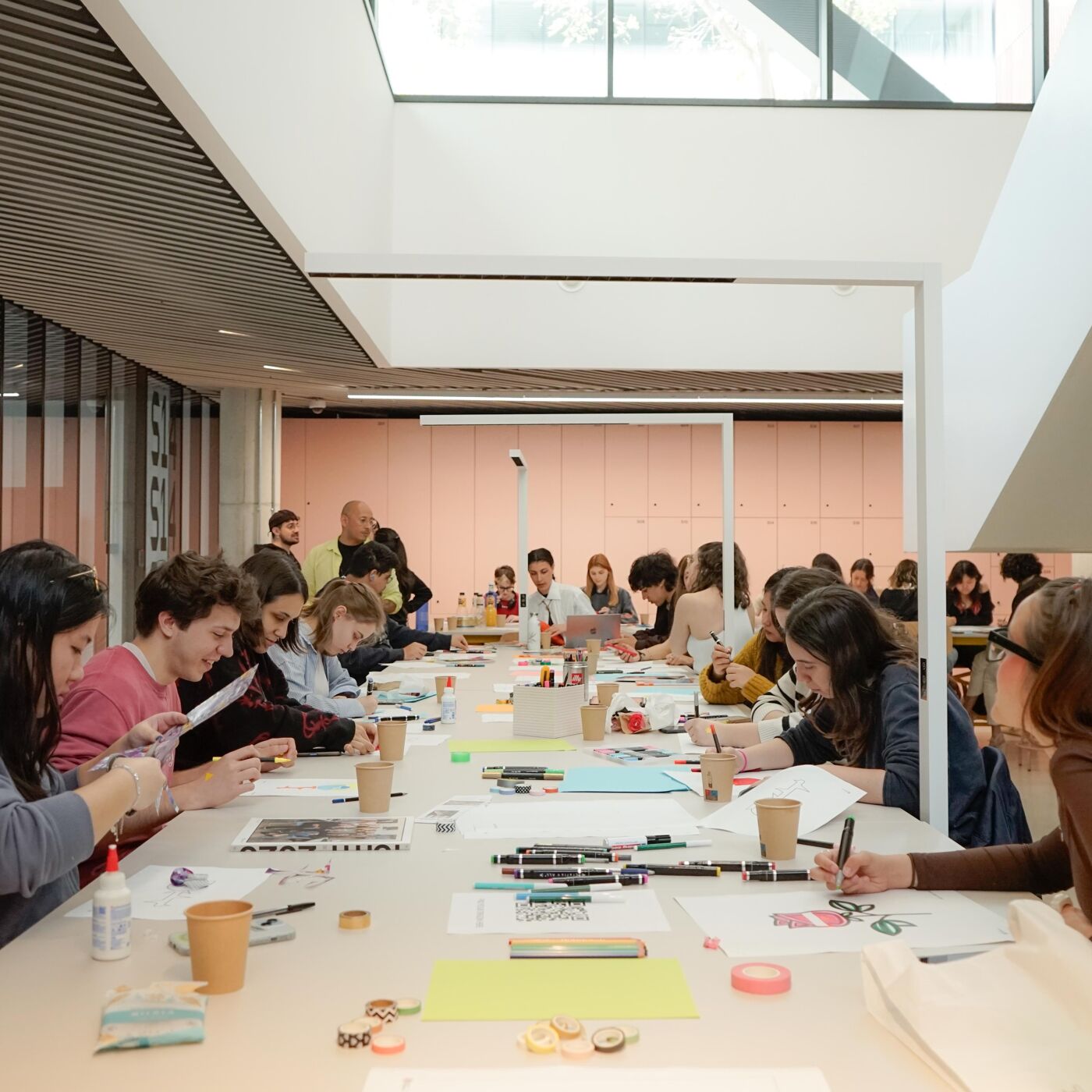 A large group of people sit at long tables in a bright, modern building, working on various projects.