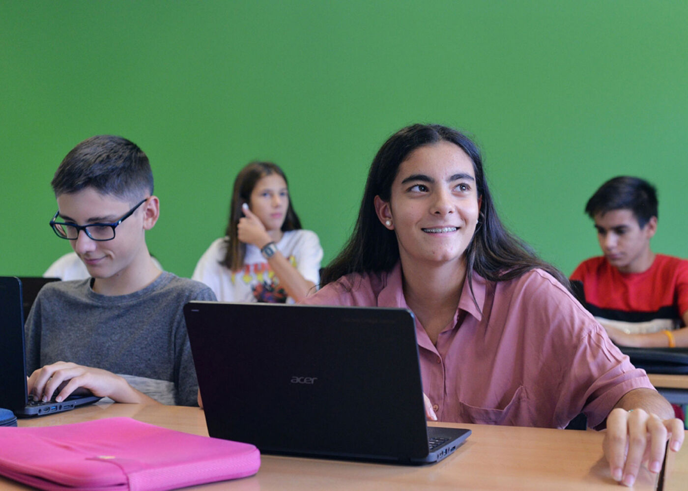 The image shows four students in a classroom setting, likely engaged in a lesson involving laptops. The girl in the foreground is smiling, while the others are focused on their screens. The background features a green wall.