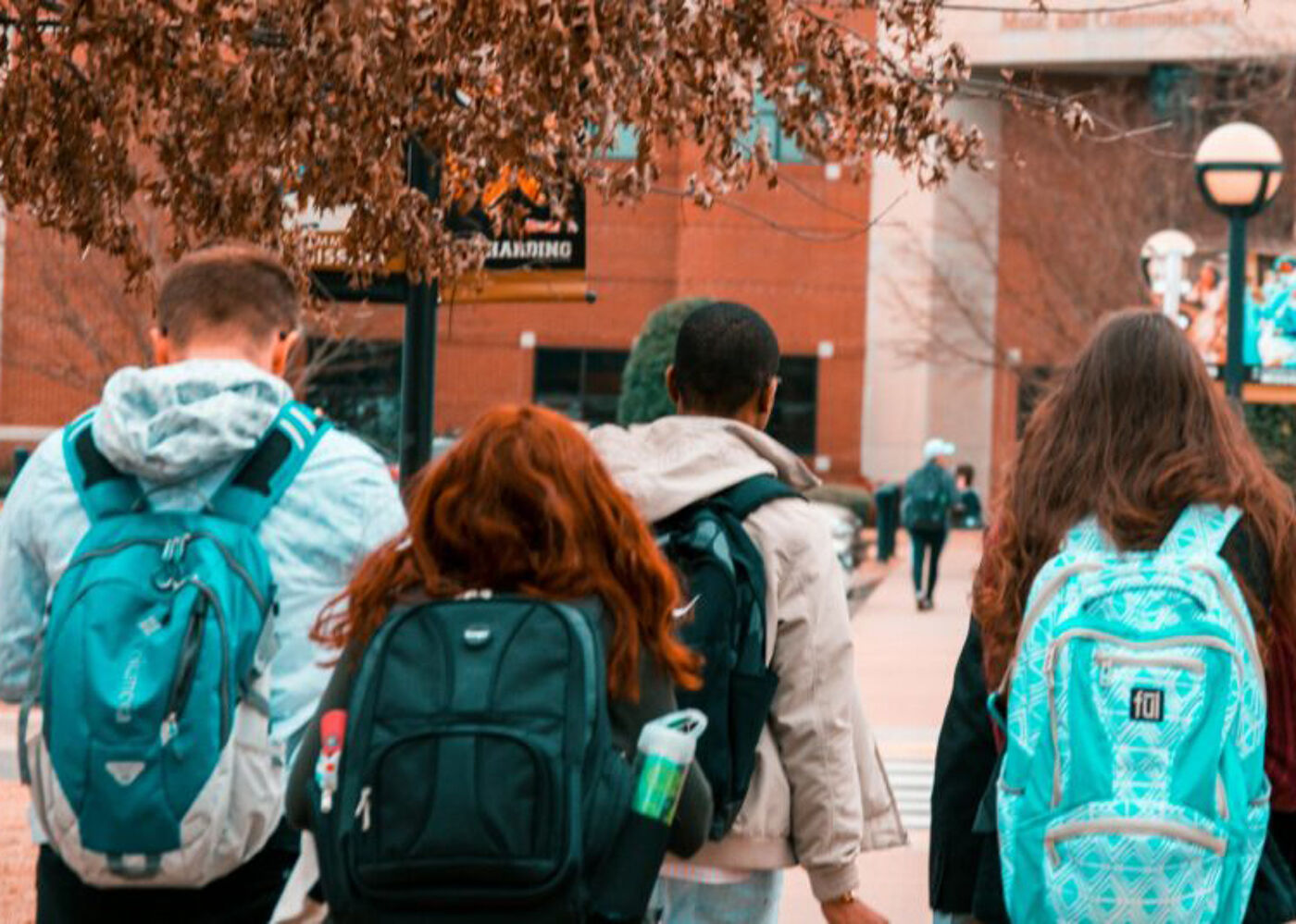 The image shows a group of college students walking on campus. They are all wearing backpacks and appear to be headed to class. The photo is taken from behind, focusing on their backs and backpacks.