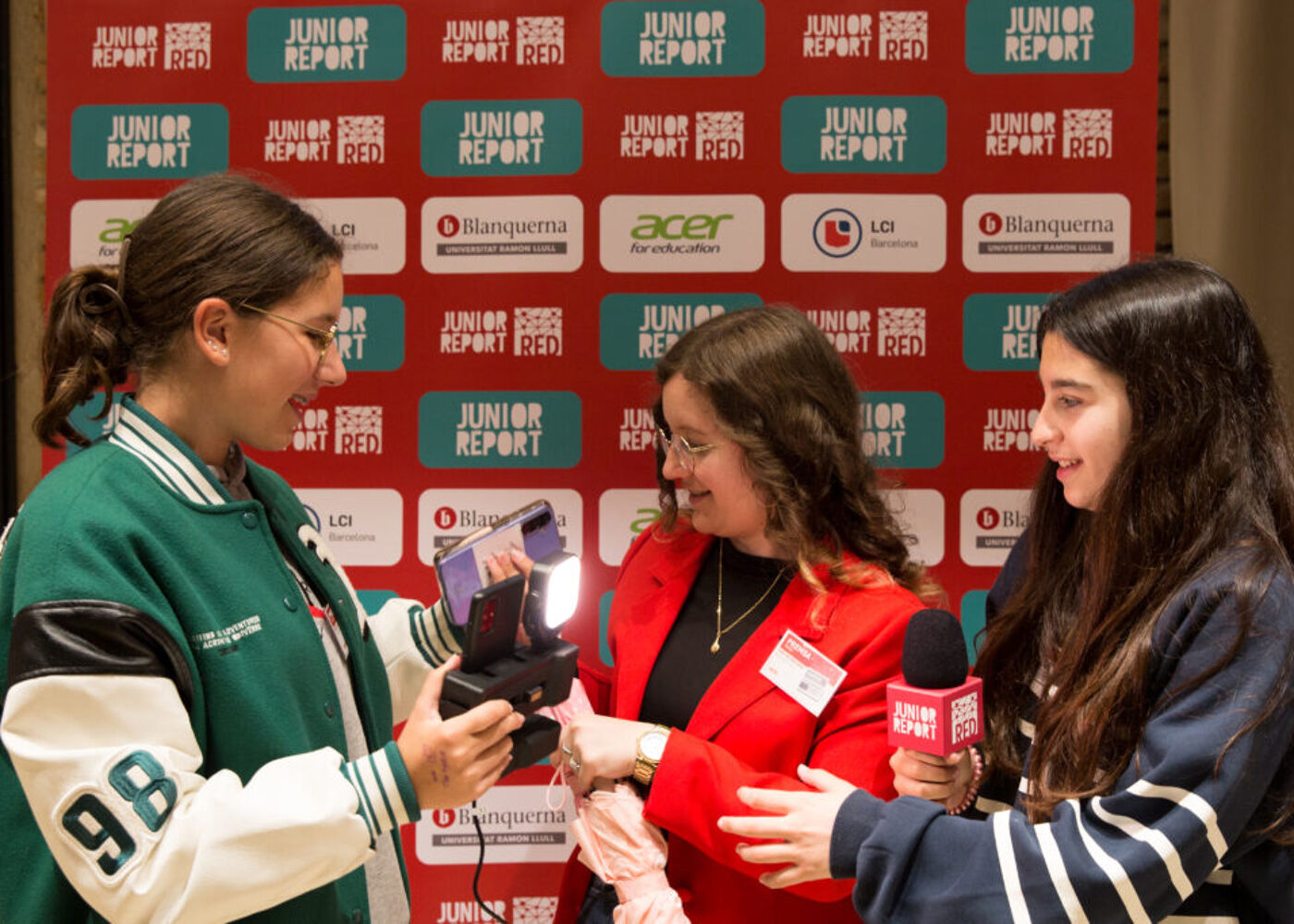 Three women are gathered in front of a backdrop with the words "Junior Report" repeated across it. One woman is holding a camera, another is holding a microphone, and the third is in the middle, possibly being interviewed or photographed.