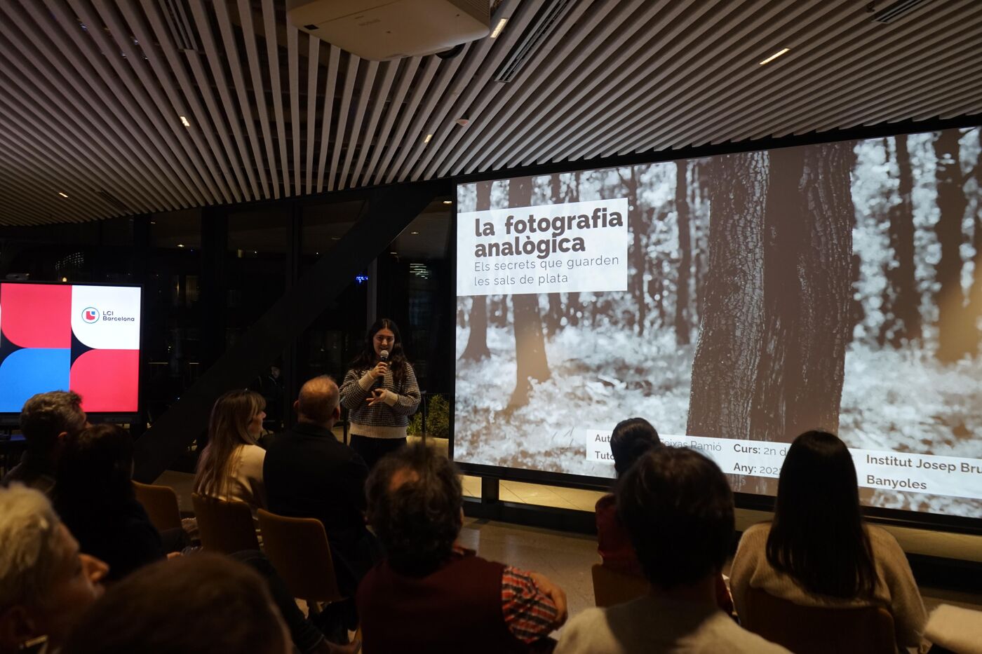 A person is giving a presentation on analog photography to an audience. The presentation screen shows a black and white image of trees in a forest. The room appears to be a modern auditorium.