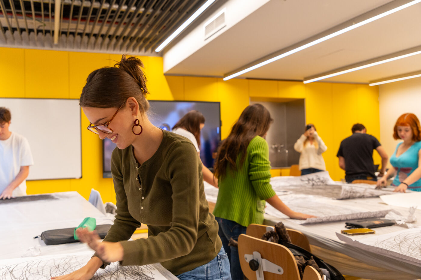 In a brightly lit room, several people are gathered around tables covered with fabric. A woman in the foreground, wearing glasses and a green shirt, is focused on her work, while others are similarly engaged in sewing or crafting activities.