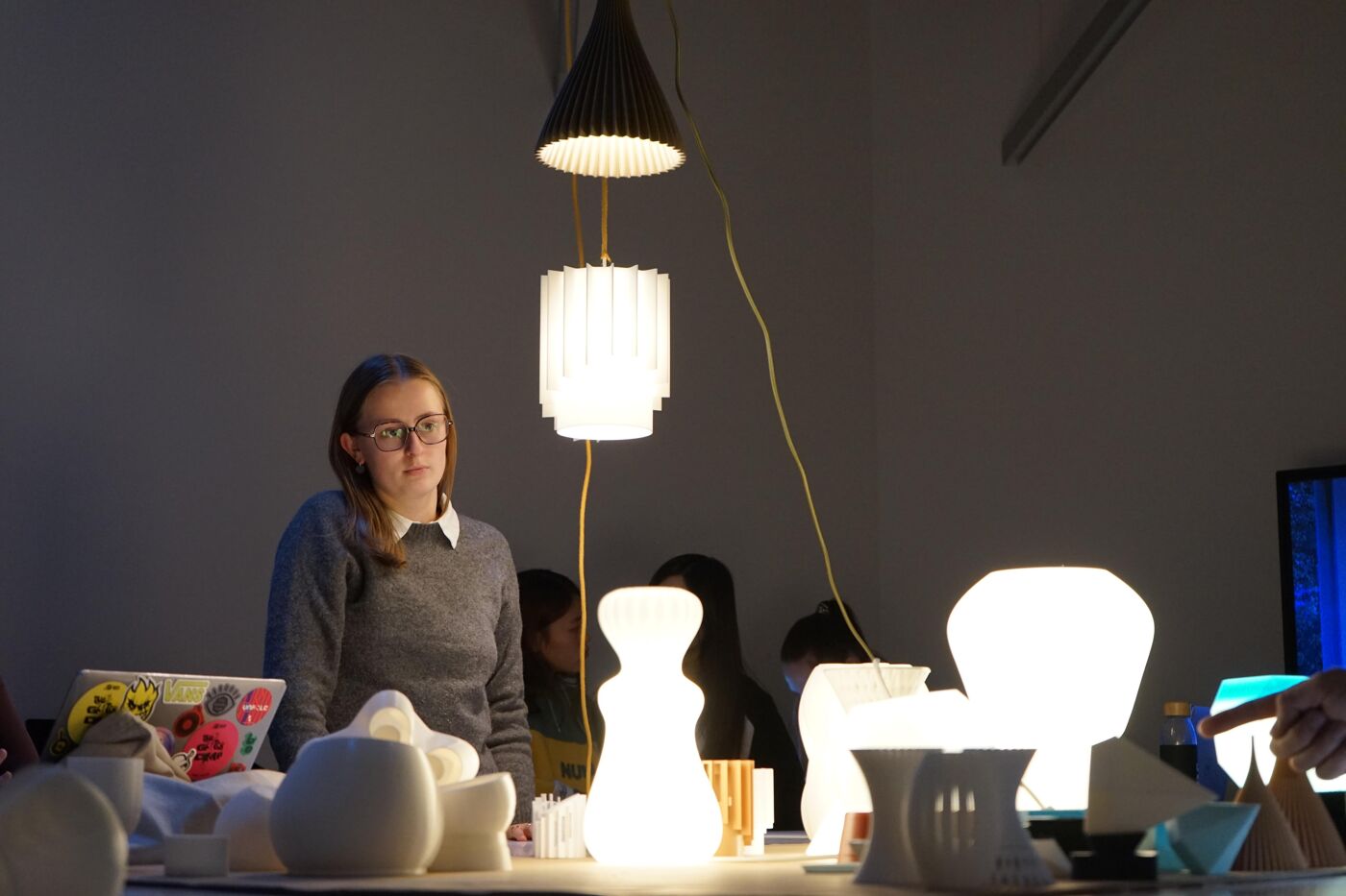 A young woman with glasses stands in front of a display of white lamps and sculptures, illuminated by various light sources.