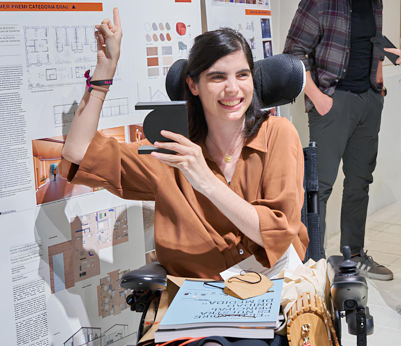 A woman in a wheelchair holds an award, smiling at the camera, surrounded by architectural designs and a man standing in the background.