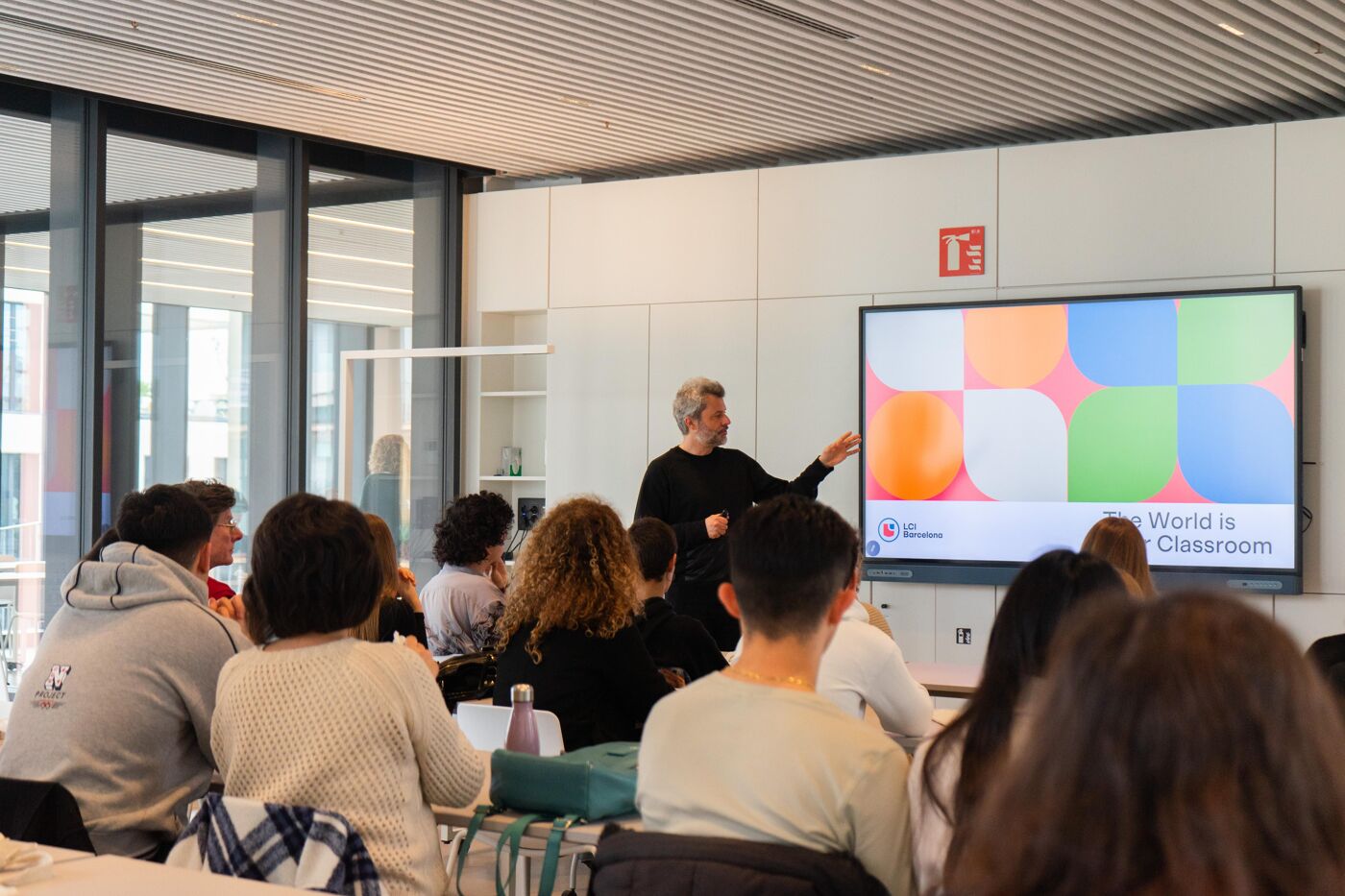 A man stands in front of a screen presenting to a group of people seated in a room with large windows.