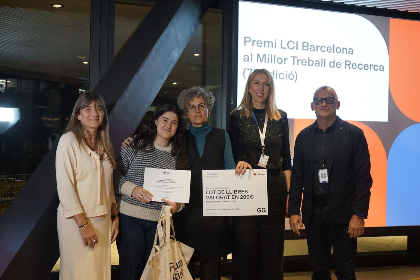 Four women and a man stand in front of a large screen displaying an award ceremony announcement, with two women holding certificates and a tote bag.