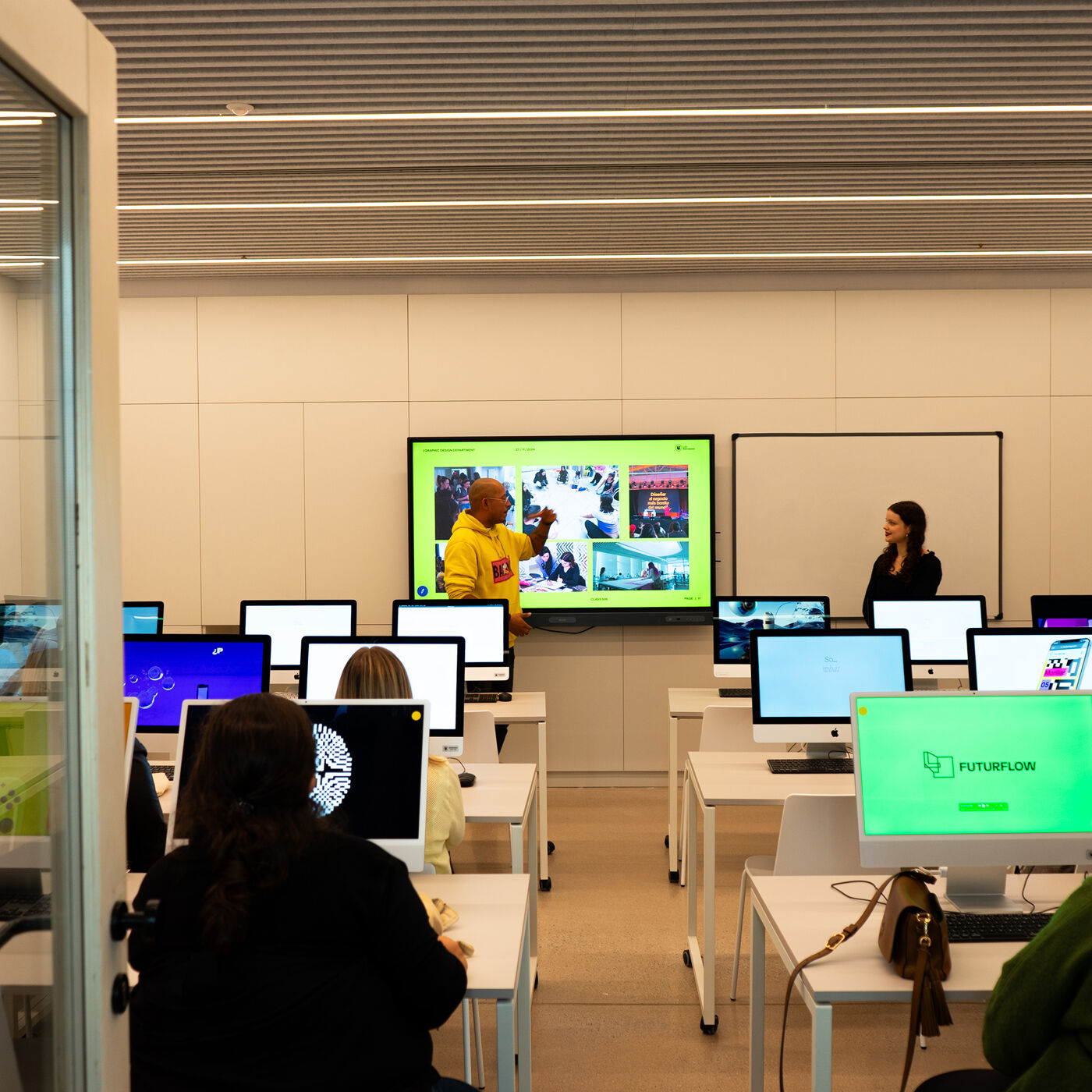 A classroom with students seated at computers, listening to a man giving a presentation on a large screen while a woman stands nearby.