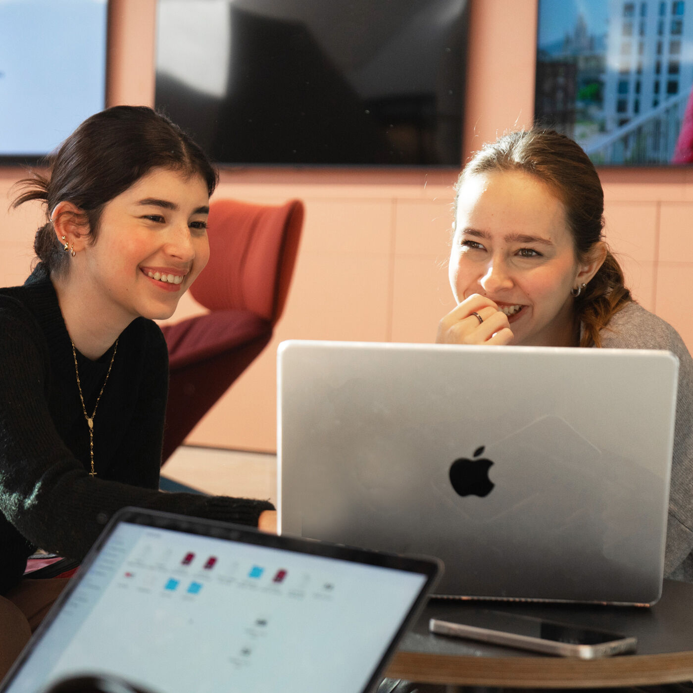 Two young women sit at a table, smiling and looking at a laptop together.