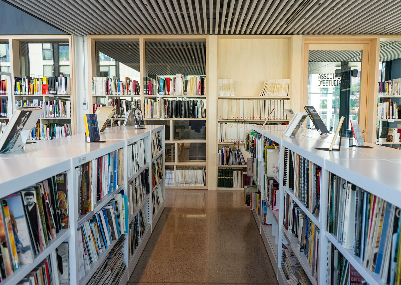 Two long white shelving units filled with books line a polished concrete floor in a modern library, with glass-fronted wooden bookcases in the background.