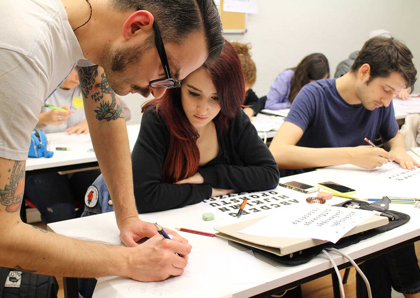 A man with tattoos is teaching a group of students calligraphy in a classroom.