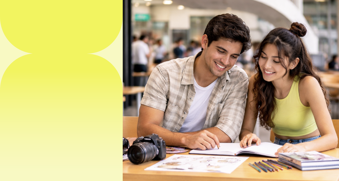 A young, smiling couple studies together in a cafe, with books, colored pencils, and a camera on the table.