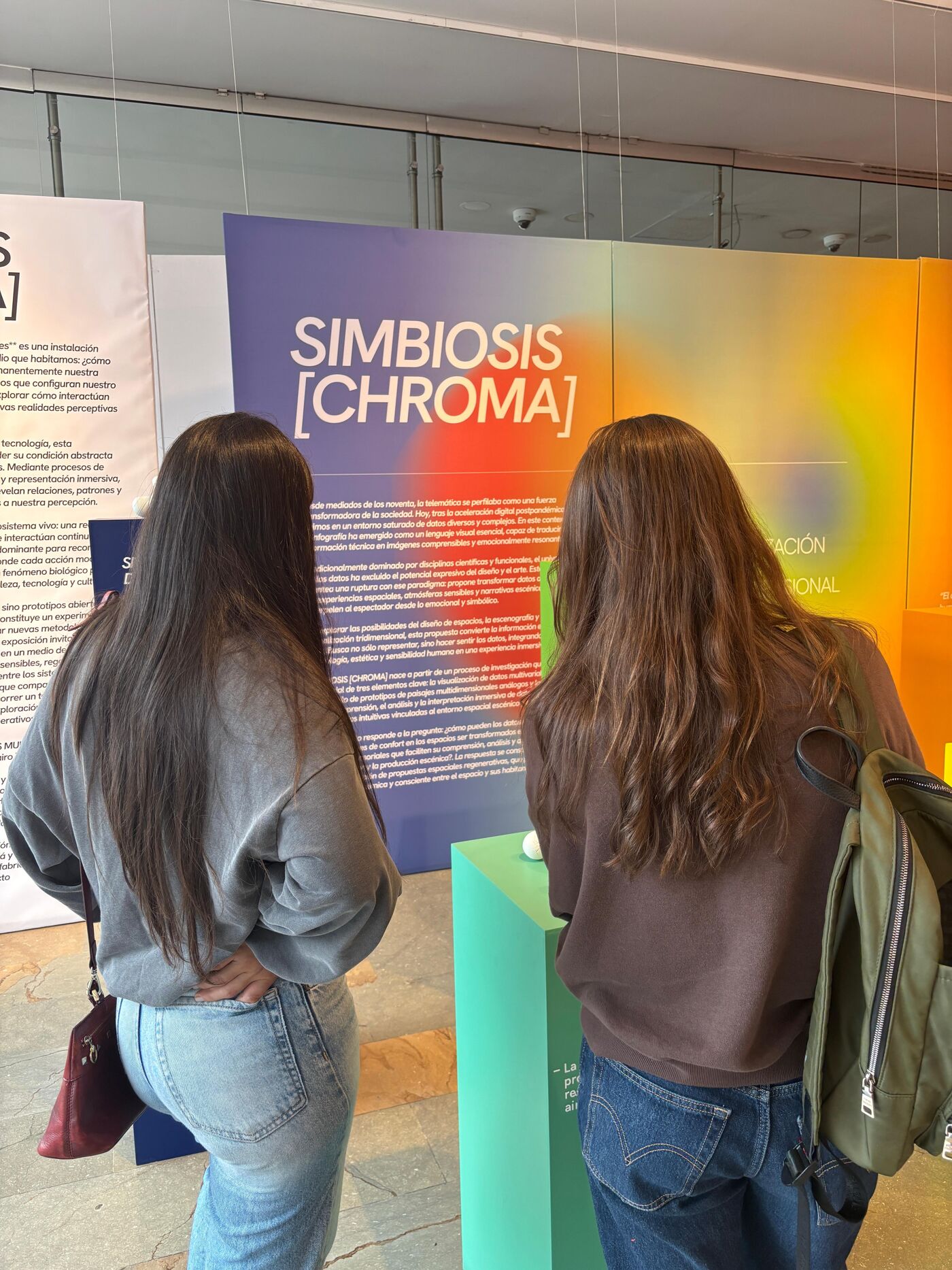 Two individuals are observing an exhibit titled "SIMBIOSIS [CHROMA]", which appears to be an artistic or scientific display exploring the concept of symbiosis and color.