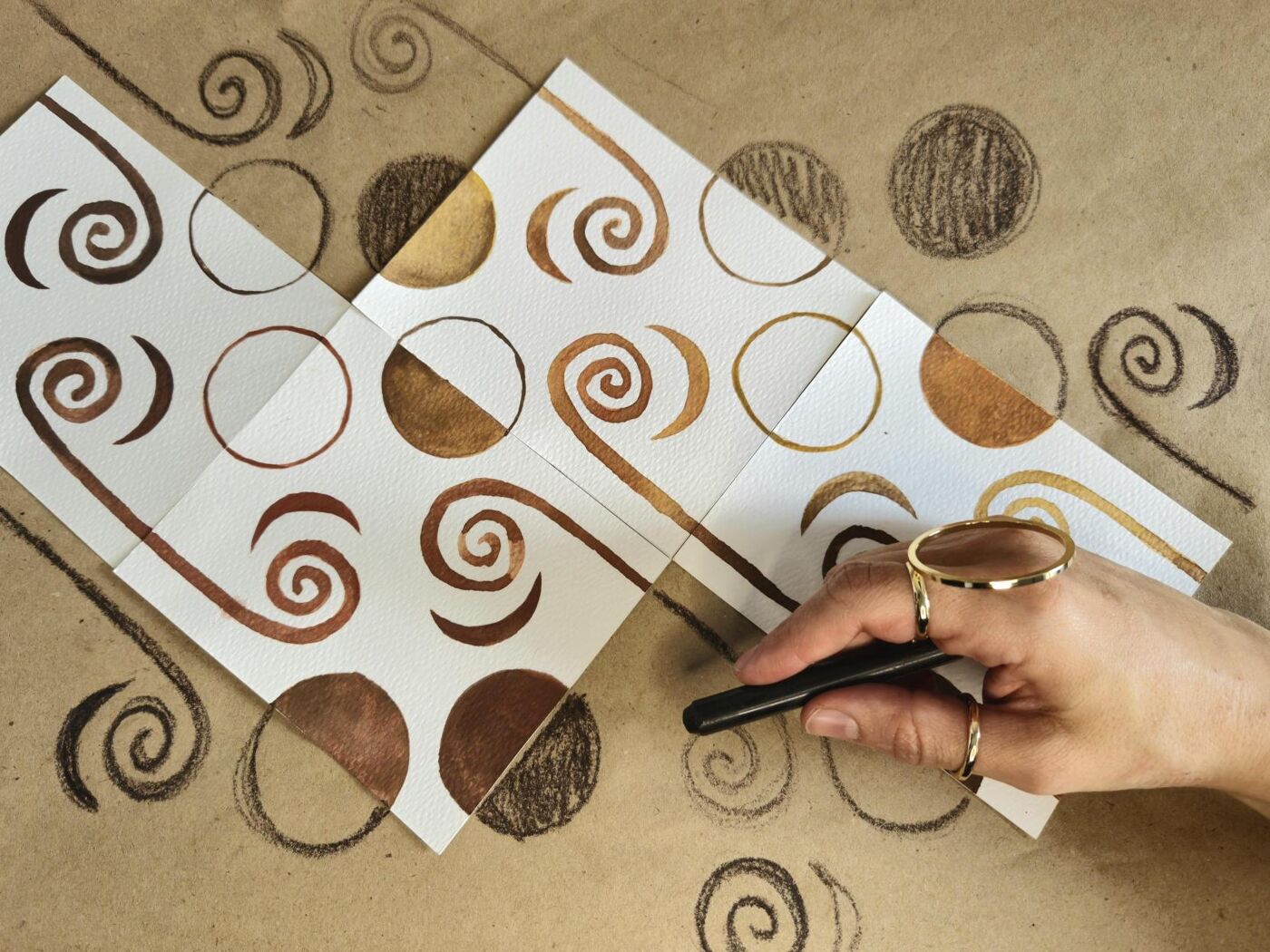 A person's hand, adorned with gold rings, holds a drawing utensil above several small watercolor paintings featuring brown spirals and circles on a beige background.