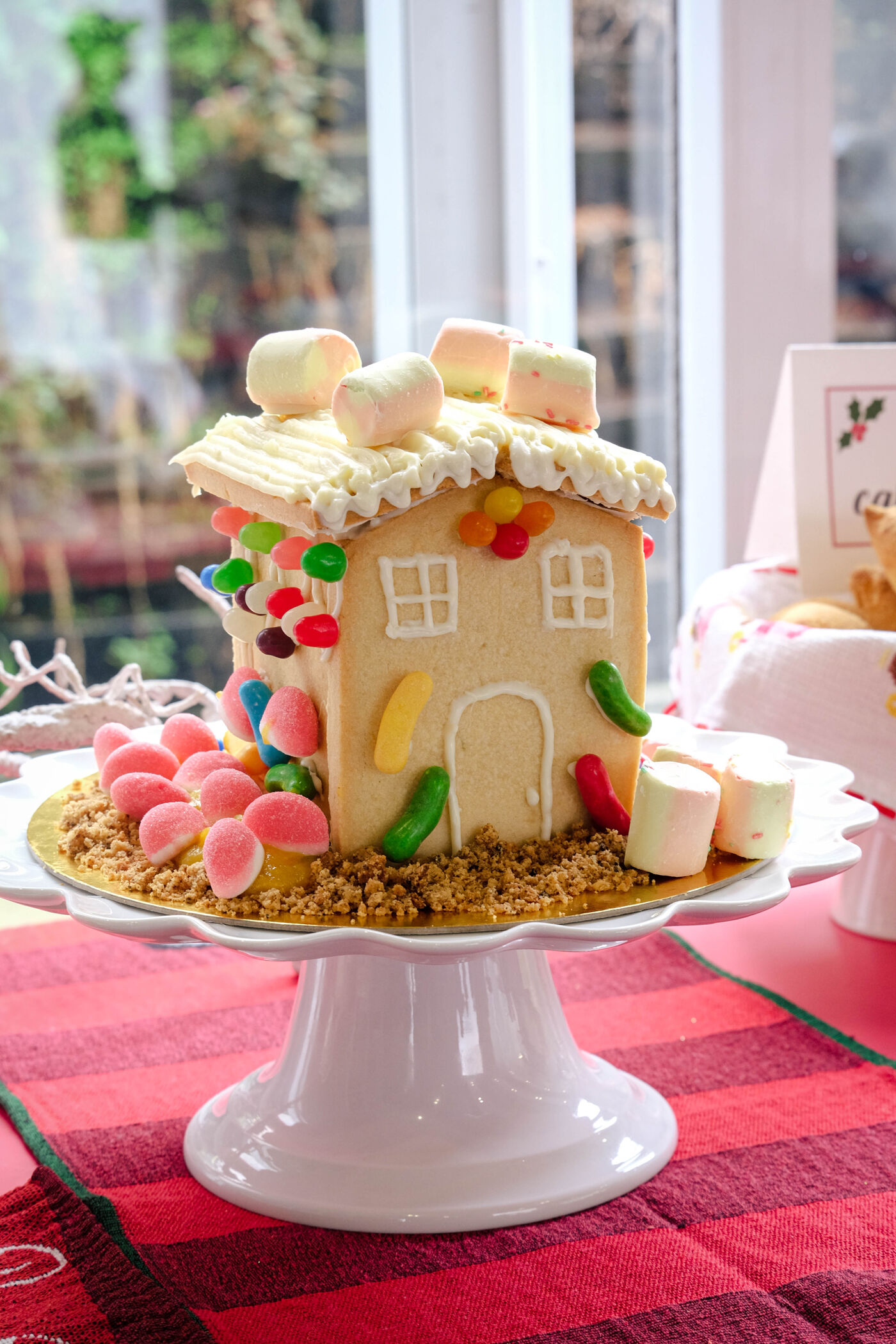 The image showcases a festive gingerbread house adorned with colorful candies, marshmallows, and a cookie roof, presented on a white cake stand.