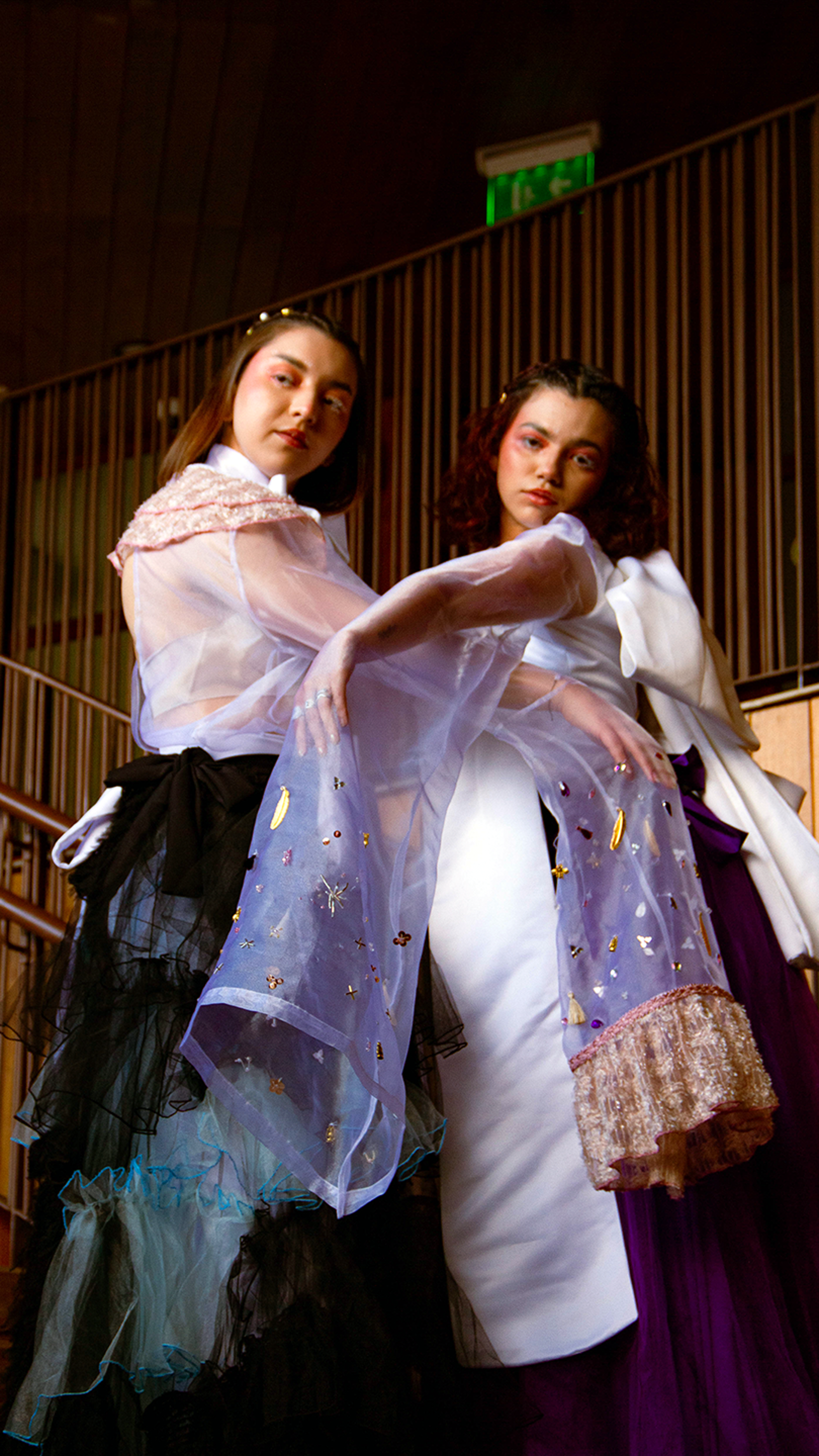 Two models poised confidently in eclectic high-fashion ensembles, exhibiting contrasting fabrics and textures against a wooden paneled backdrop.