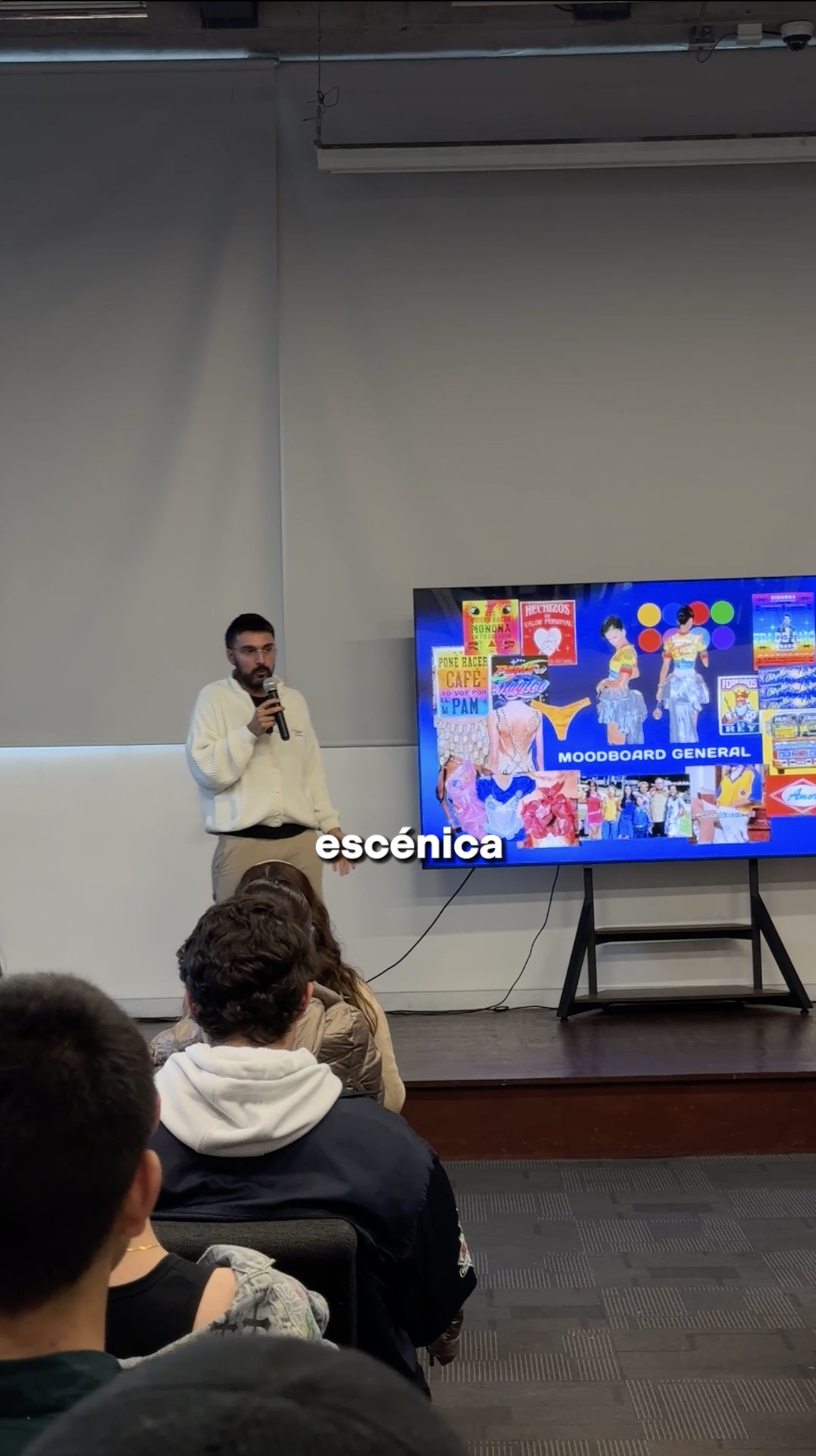 A man is giving a presentation in front of an audience. He is standing next to a large screen displaying colorful images and text. The audience is seated and listening attentively. The room appears to be a lecture hall or conference room.