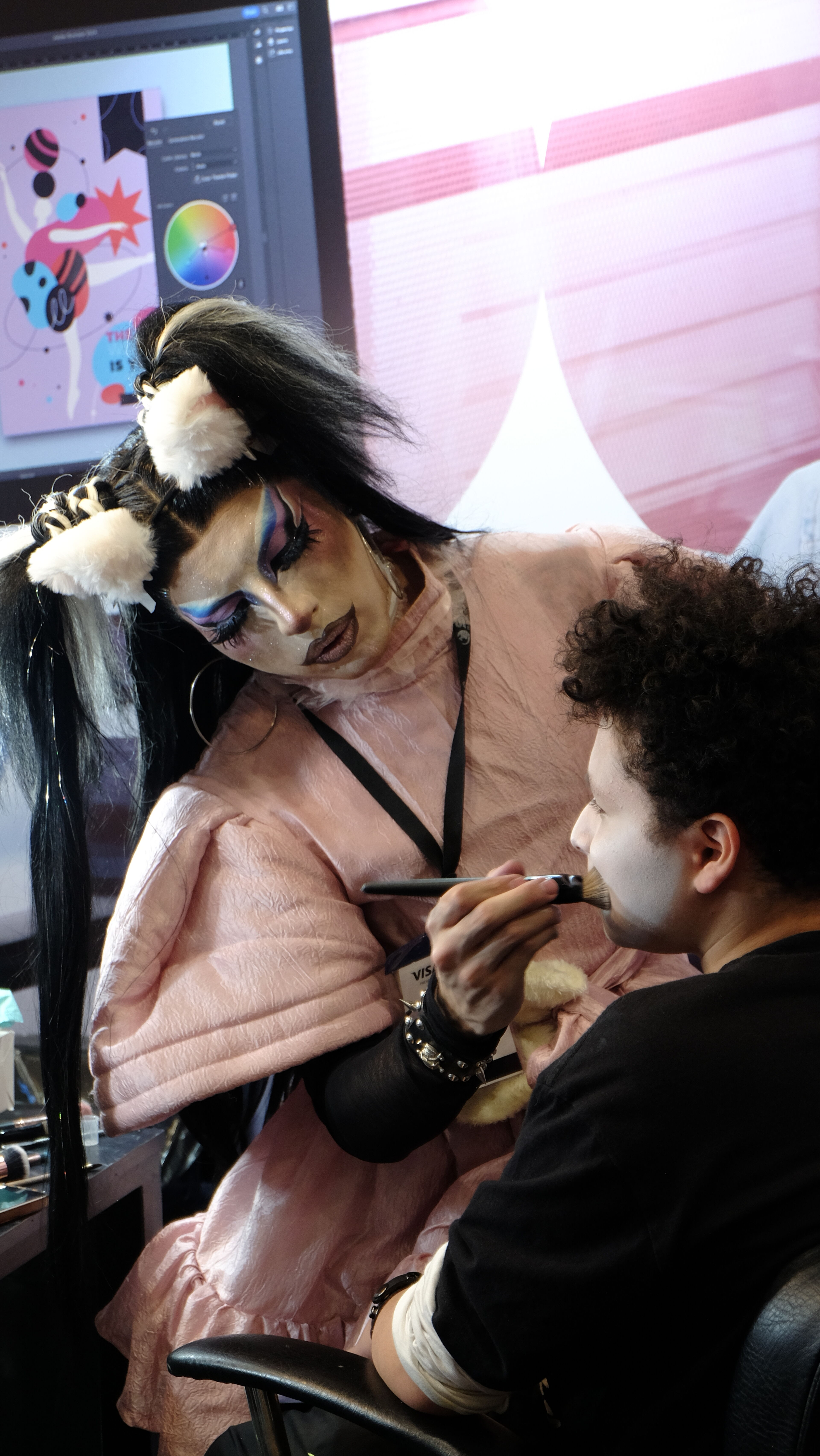 A makeup artist in dramatic attire applies intricate face paint to a client. The artist wears a pink outfit and elaborate eye makeup, creating a visually bold look in a lively, artistic environment.