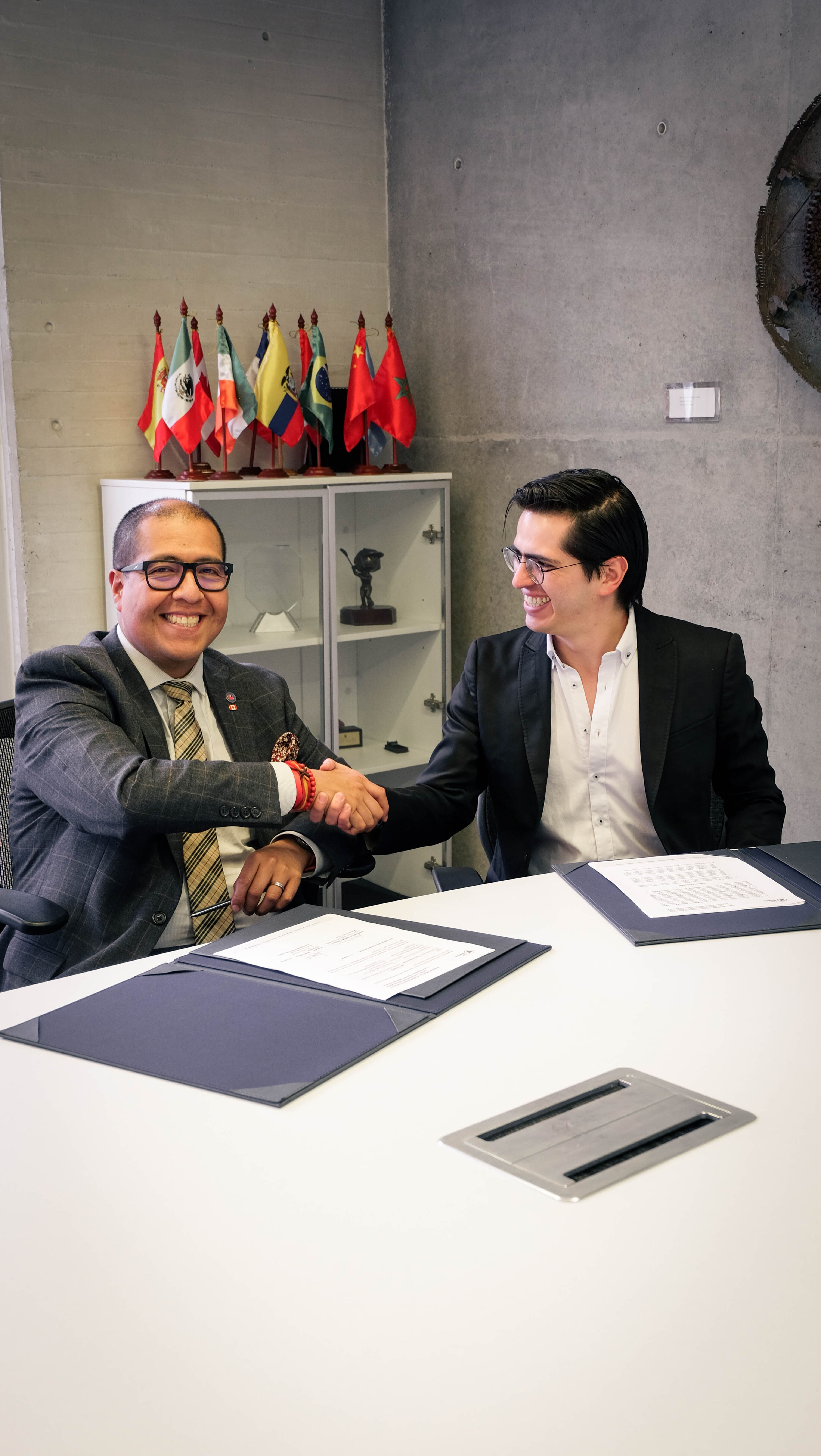  Two men in formal attire shake hands over signed documents, symbolizing a partnership agreement. They smile warmly in a professional setting adorned with various international flags in the background.