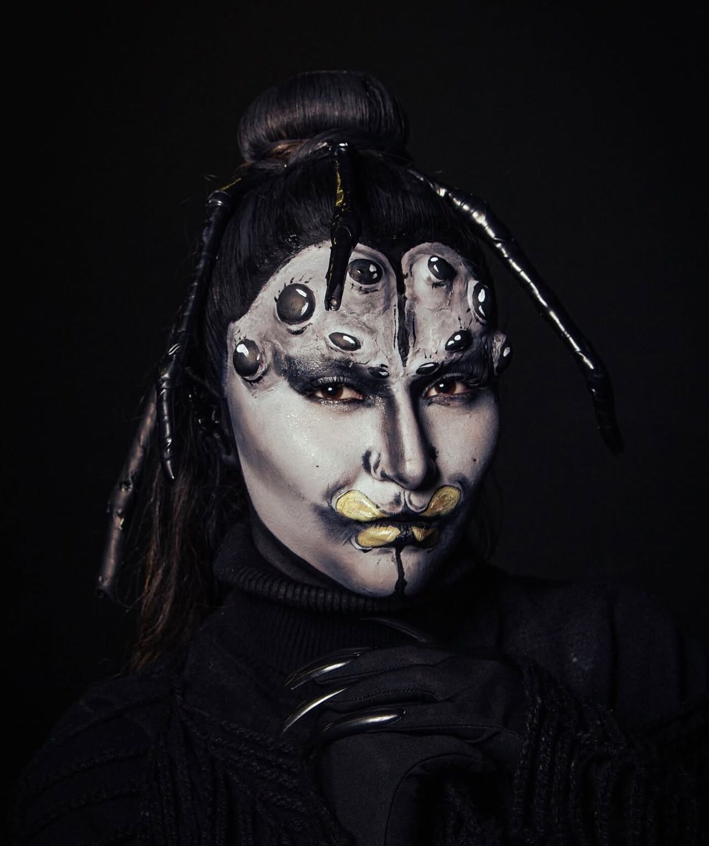 The image shows a person with elaborate spider-themed makeup. Their face is painted in grayscale with multiple eyes added to the forehead. The lips are highlighted with a yellow-green color, and spider-like appendages extend from the head.