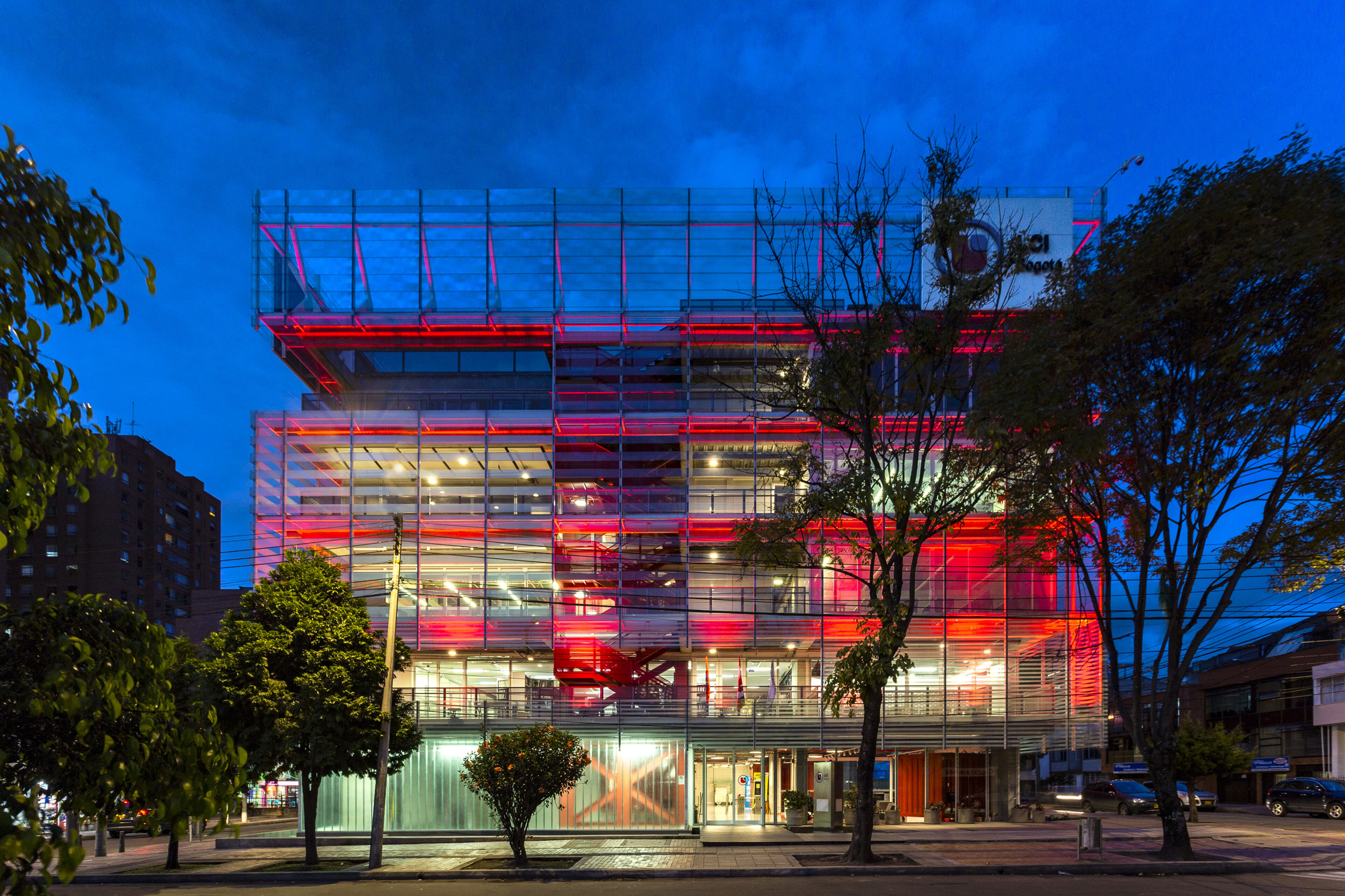 A contemporary building bathed in vibrant red lighting stands out against the evening sky, its glass facade reflecting the ambient city light.