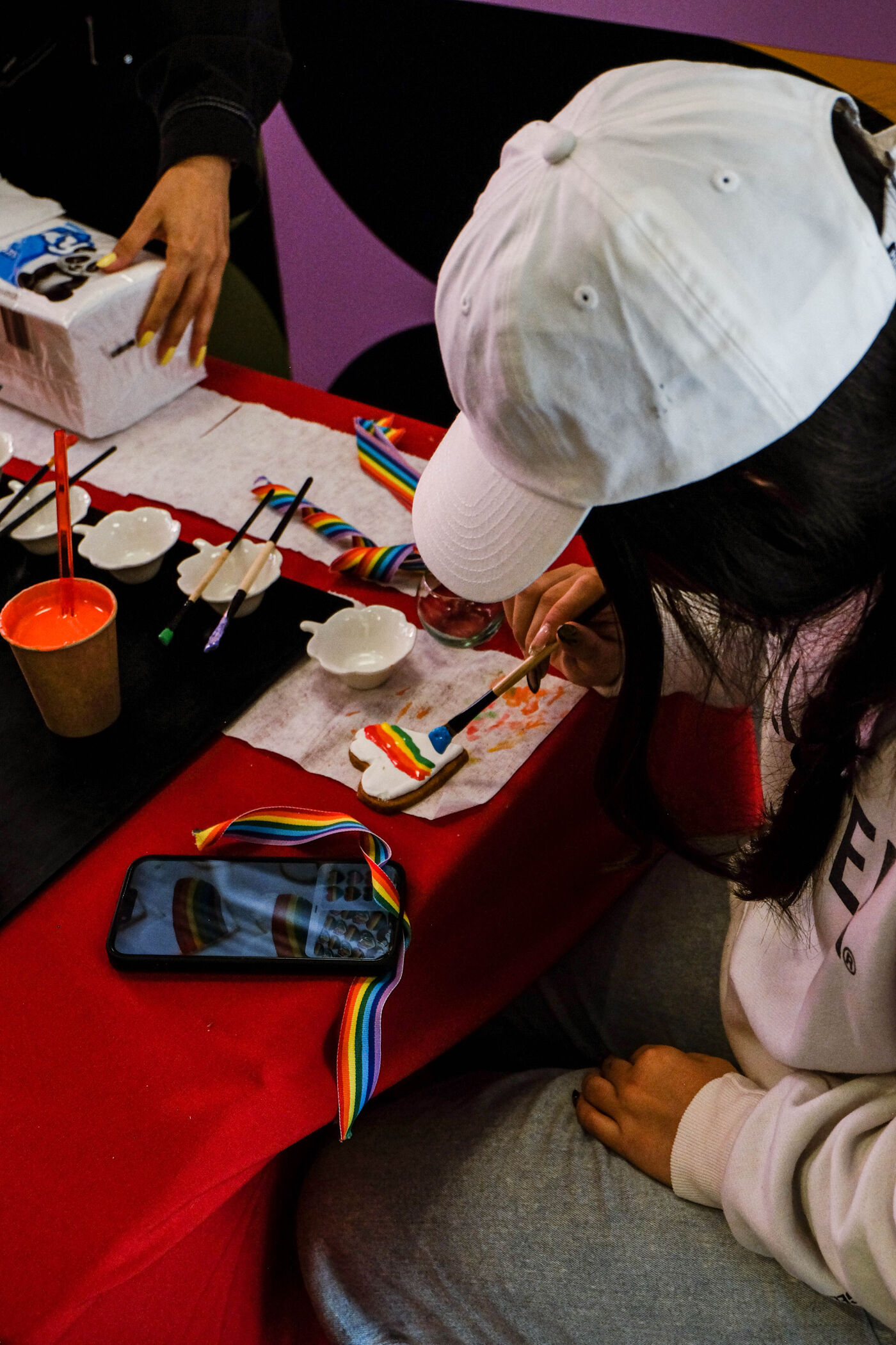 A person wearing a white cap is painting a rainbow on a small object at a red table. There are paint containers, brushes, and a phone with a rainbow image on the table.