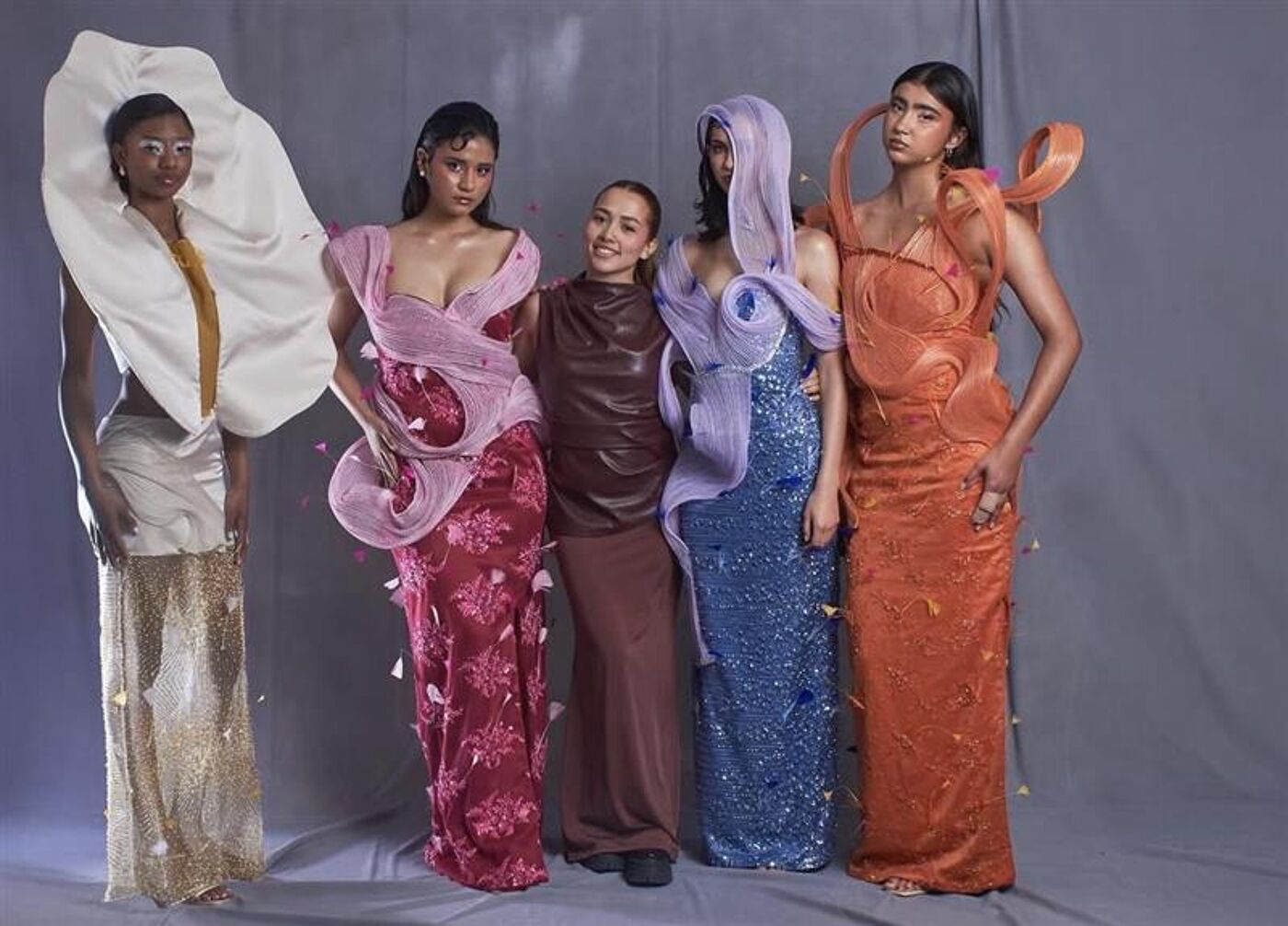 Five women are posing in a studio. They are wearing colorful and unique dresses. The dresses appear to be made of unconventional materials and have avant-garde designs. The background is a plain gray.